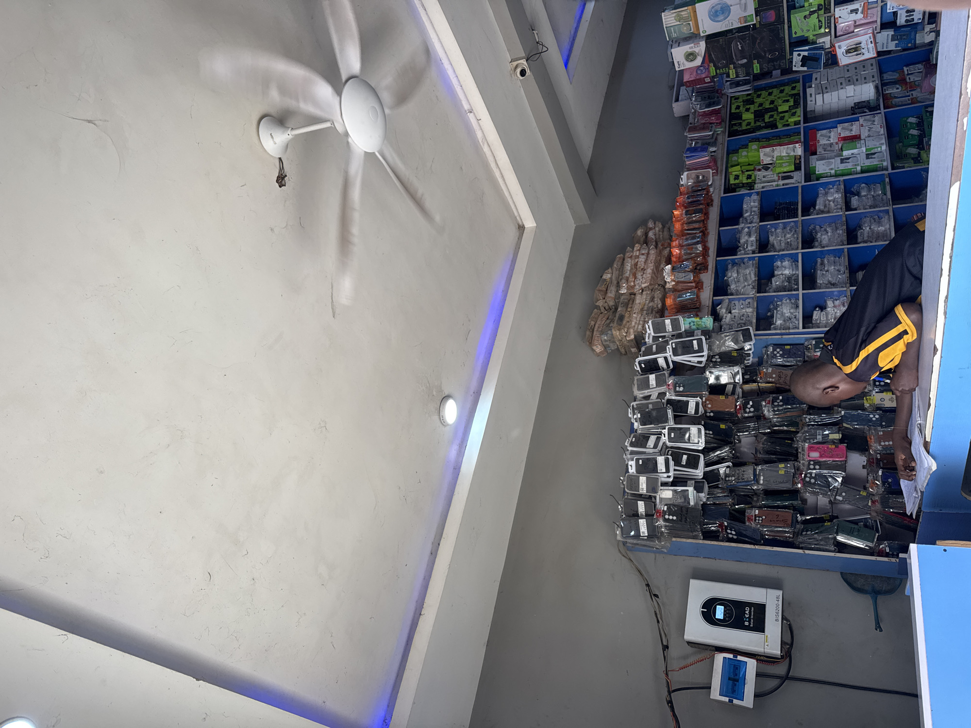 The image depicts a small electronics shop in Jalingo, Nigeria. A young man, wearing a black and yellow shirt, is seen from behind, looking down at papers on a counter. He appears to be an employee or owner of the shop. The shop's interior is lined with shelves displaying a variety of mobile phone accessories, including numerous phone cases, chargers, and other electronic gadgets. In the background, a ceiling fan is in motion, and blue LED strip lights are installed along the ceiling's edges, creating a modern ambiance. On the left wall, a solar power inverter is mounted, suggesting the shop utilizes solar energy. The overall scene suggests a busy retail environment focused on mobile phone accessories.