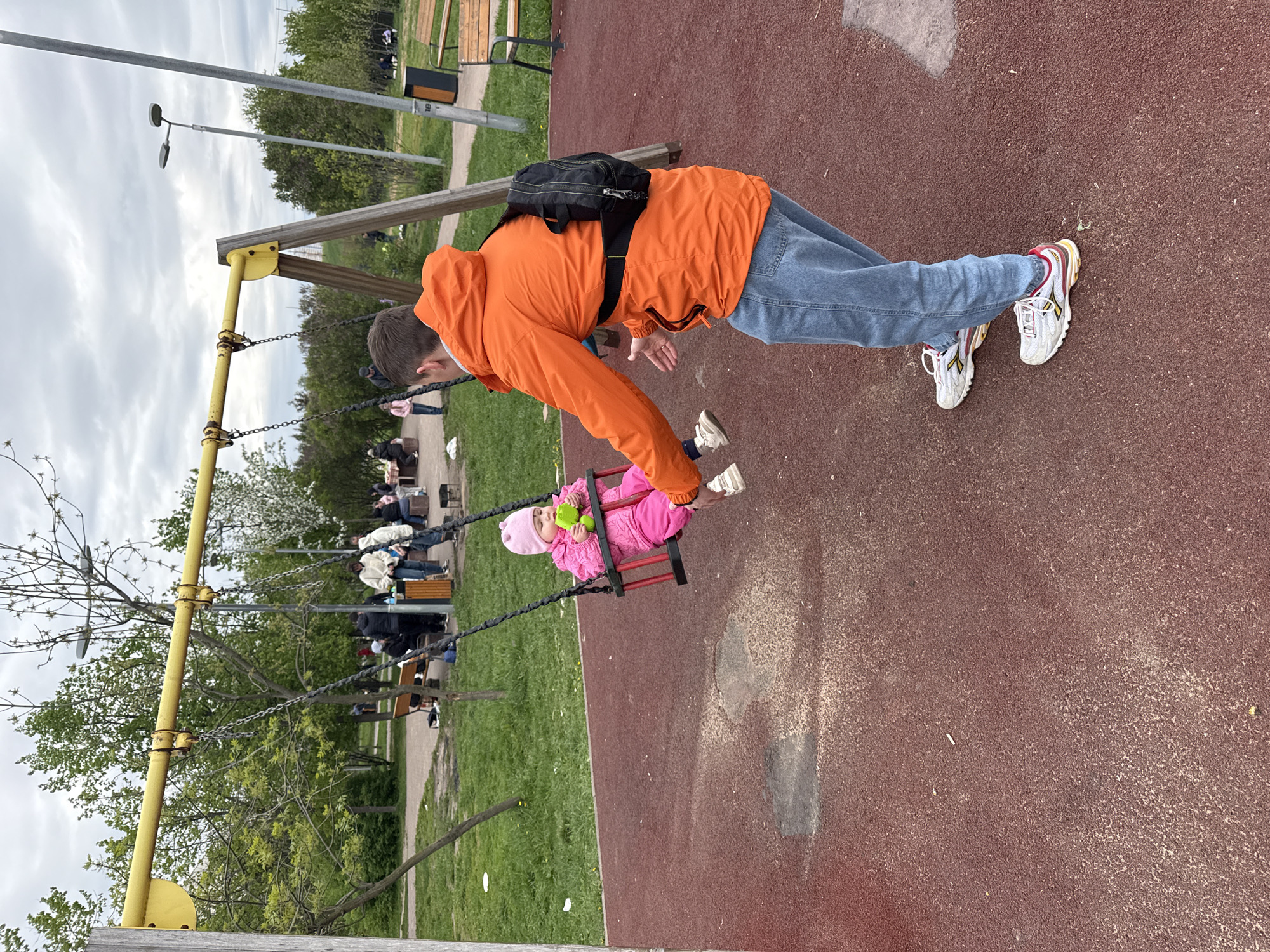 A person wearing an orange jacket, blue jeans, and white sneakers is seen pushing a child in a pink outfit and hat on a swing. The person has a black bag slung across their back. The swing set is made of yellow metal and has metal chains. The ground is covered in reddish-brown surfacing, likely in a playground. In the background, other people are sitting on benches near trees and green grass. The sky is overcast. The location is Moscow, Russia.