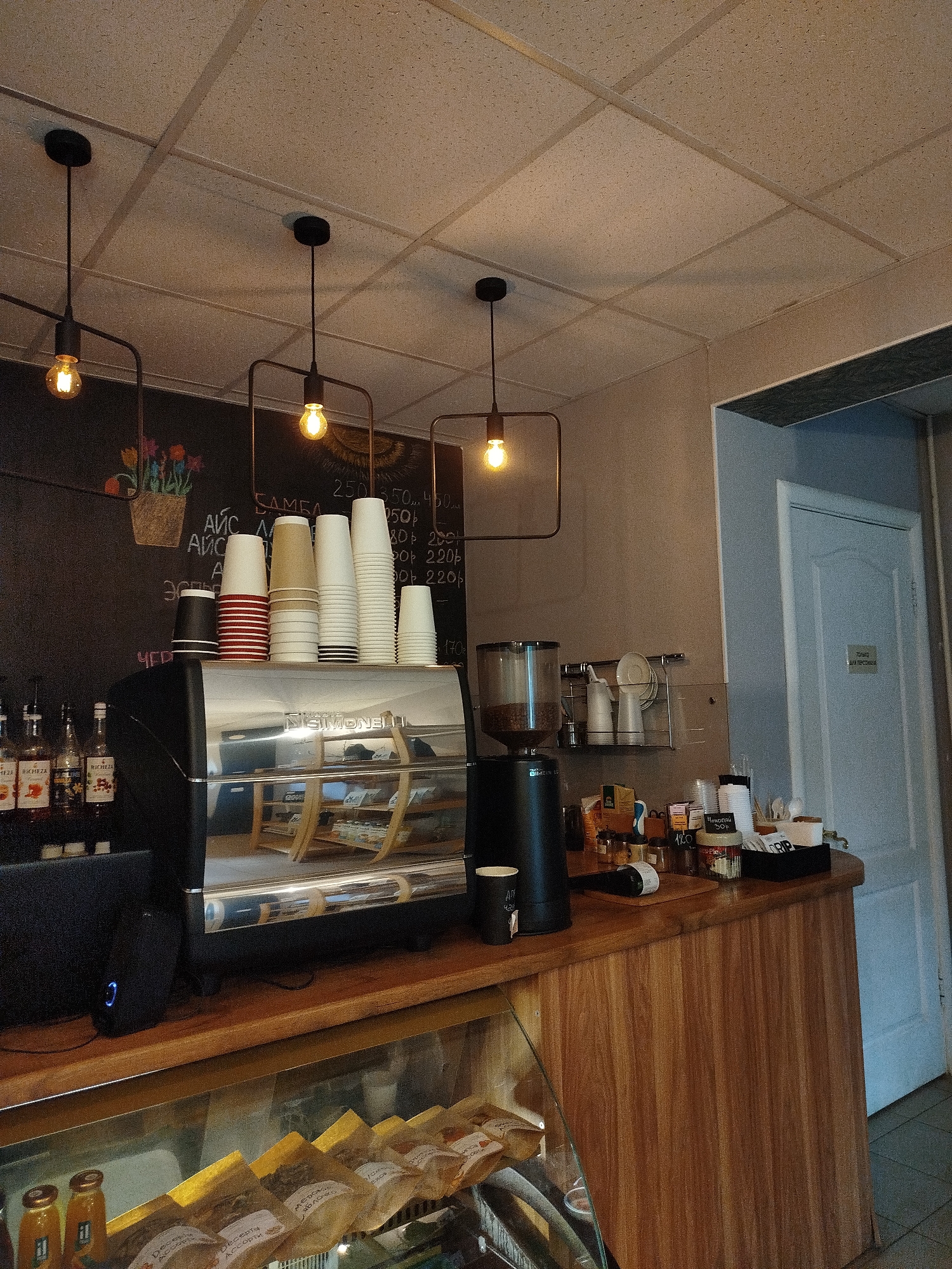 The image depicts the interior of a cafe. In the foreground, a wooden counter is visible, with a glass display case showcasing food items wrapped in paper. To the left, a coffee machine with a shiny metal surface stands prominently. Above the counter, several stacks of paper cups are arranged on a shelf, with a chalkboard wall behind them featuring drawings and handwritten text. Industrial-style pendant lights hang from the ceiling. Further back, a door is visible to the right, along with more shelving and a coffee grinder. The overall environment suggests a functional and welcoming cafe setting.