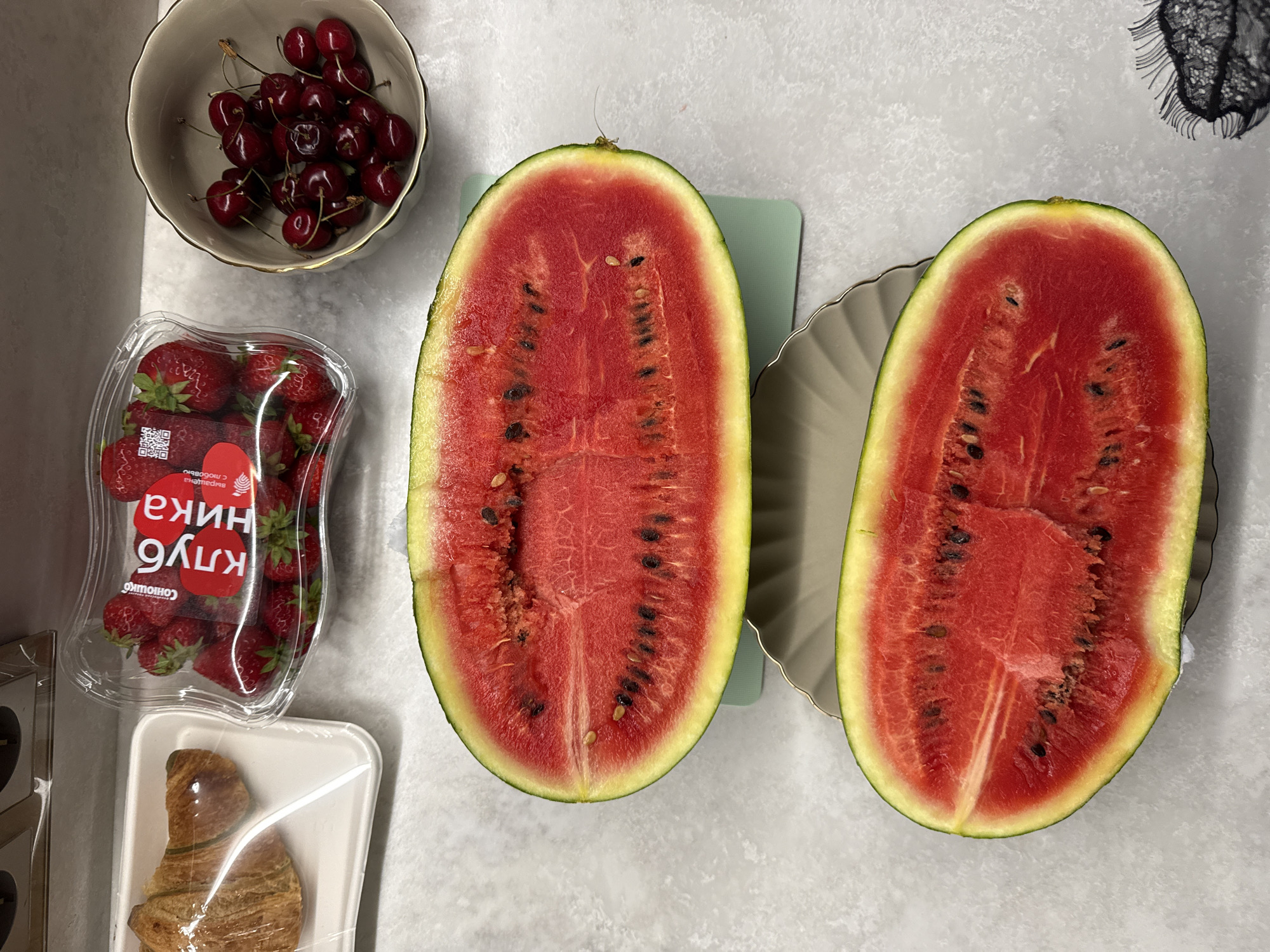In a domestic kitchen setting, likely in Khimki, Russia, a tabletop displays a variety of food items. Two halves of a cut watermelon rest on a light gray surface. The top half is positioned on a light green cutting board, while the bottom half is on a scalloped gray plate. To the left, a croissant is presented in a clear plastic container. Next to it, a clear plastic clamshell package holds a quantity of red strawberries, with Russian text visible on a red label. To the right, a small ceramic bowl contains a pile of ripe cherries. The overall scene is well-lit, with the light source originating from the top.