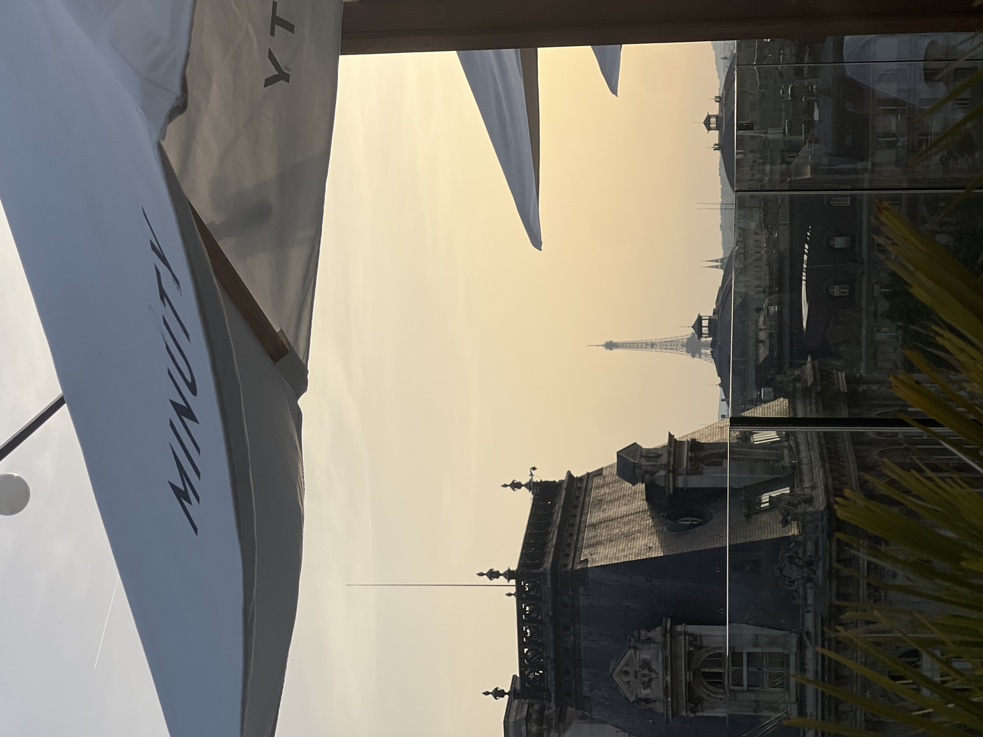 The image captures a view of Paris, France, likely taken from a rooftop or high-level terrace. The Eiffel Tower is visible in the distance, partially obscured by the hazy, sunlit sky. In the foreground, an ornate building with detailed architectural features and a slate roof is prominent. Parts of sun umbrellas with branding such as "MINUTY" are in the top of the picture. Some plants are visible on the right side of the frame. The perspective is somewhat upward, capturing the skyline and architectural details in the scene.