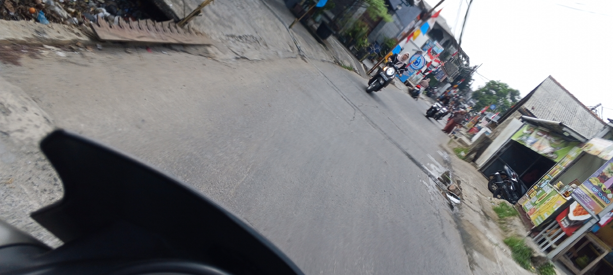 The image depicts a street scene in Depok, Indonesia, likely on a slightly inclined road. The foreground shows the dark dashboard or windshield of a vehicle, with the rest of the image capturing the street ahead. Several motorcycles are present, some moving and some parked. In the middle ground, a dark-colored scooter with two individuals, a driver and a passenger wearing headscarves, is riding on the road. Further down the street, more motorcycles are visible, along with several small shops or stalls lining the right side of the road, some displaying colorful signs with text in Indonesian. One stall has a price list indicating "Rp 10.000". The environment appears to be a typical urban street with buildings, some greenery, and evidence of trash on the left side of the road. The overall atmosphere suggests a busy commercial and residential area.