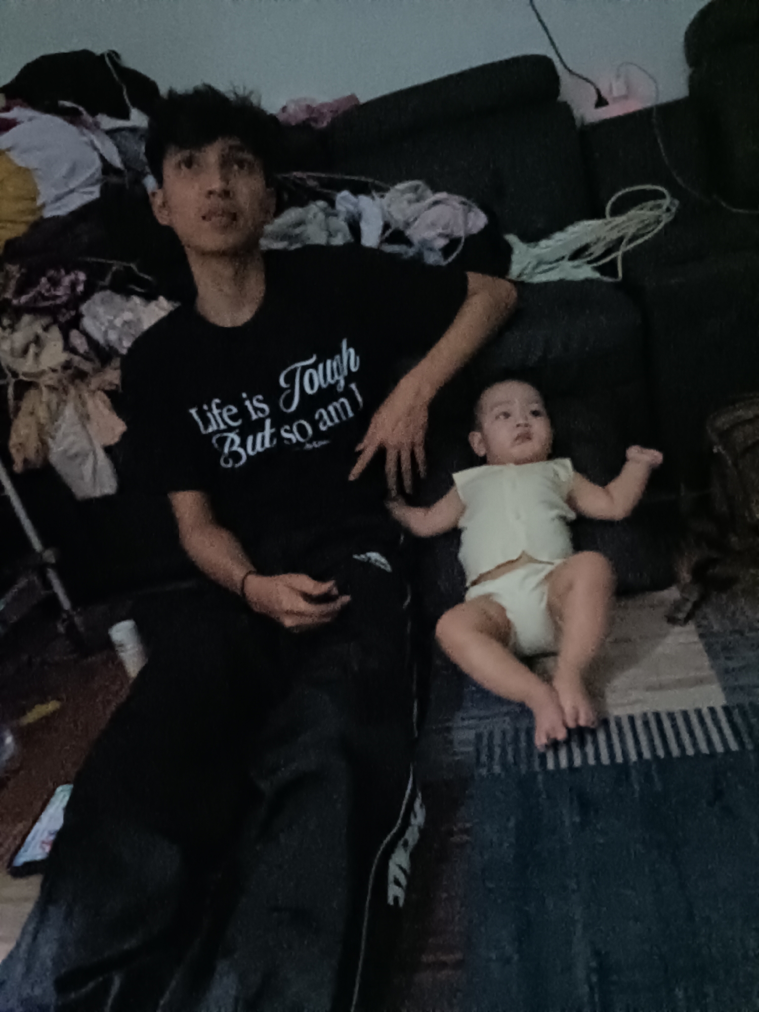 A young man is sitting on a couch with a baby on his lap, indoors. The man is wearing a black t-shirt with white text that reads "Life is Tough But so am I". He has short, dark hair and is looking to his left. The baby is wearing a light-colored onesie and is looking forward with its arms raised. The couch is dark and appears to be in a living room. There are various items of clothing and fabric scattered around the room, including on the couch and on the floor. The lighting in the room is dim, suggesting it may be evening or nighttime. The scene is likely a residential interior in West Jakarta, Indonesia, based on the provided context, though specific location details are not visually apparent.