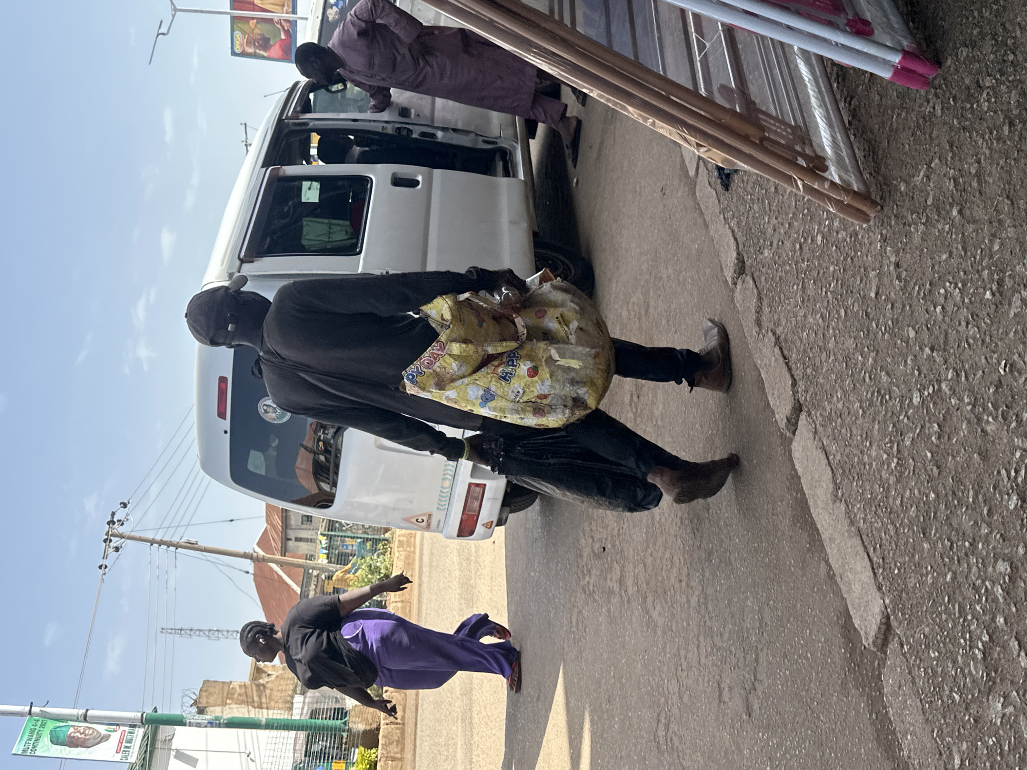 In the city of Bukuru, Nigeria, a street scene unfolds under a bright sky. A white van, its side door open, is parked on a paved road. A person wearing a dark long-sleeved shirt and dark pants, with a dark cap covering their head, walks away from the van, carrying a large, light-colored bag in their left hand and a black bag in their right. Their feet are visible, one bare and the other in a brown shoe. To the right of the van, another person in a dark mauve robe and pants stands with their hands on their hips, looking towards the van. Further left, a person in a black t-shirt and purple pants walks away from the camera. In the background, buildings and utility poles are visible, with a banner featuring a portrait and text attached to a pole. Metal shelving units are propped up on the right side of the foreground. The ground is a mix of asphalt and rougher textured surfaces.