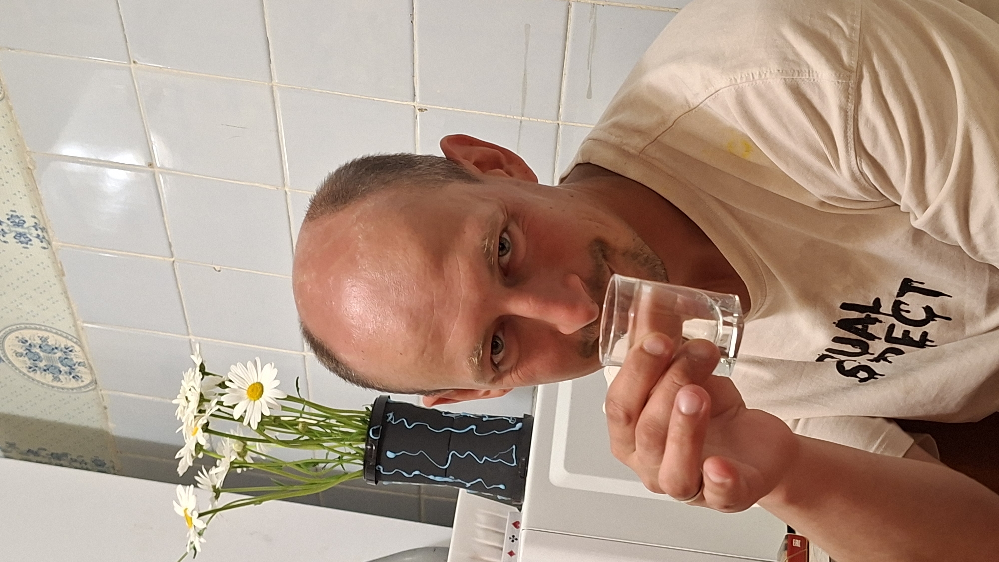 A man is holding a small glass in a bathroom. He has short brown hair and is wearing a beige t-shirt with black text. Next to him is a black vase decorated with blue wavy lines, containing daisies. The bathroom has white tiles on the walls and blue floral-patterned tiles in a border. The location is Забалоцкі сельскі Савет, Belarus.