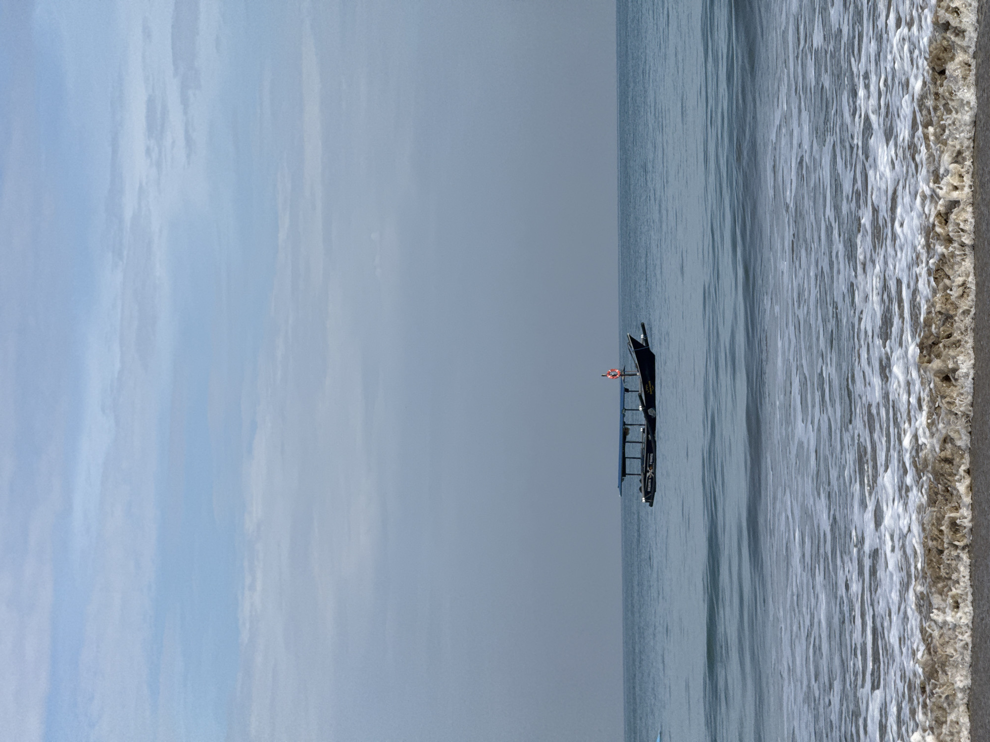 The scene is a beach near the city of Kuta Selatan, Indonesia. A black boat with a red life preserver is on the water, which is calm, with small waves close to shore. The beach sand is dark. The sky is overcast.