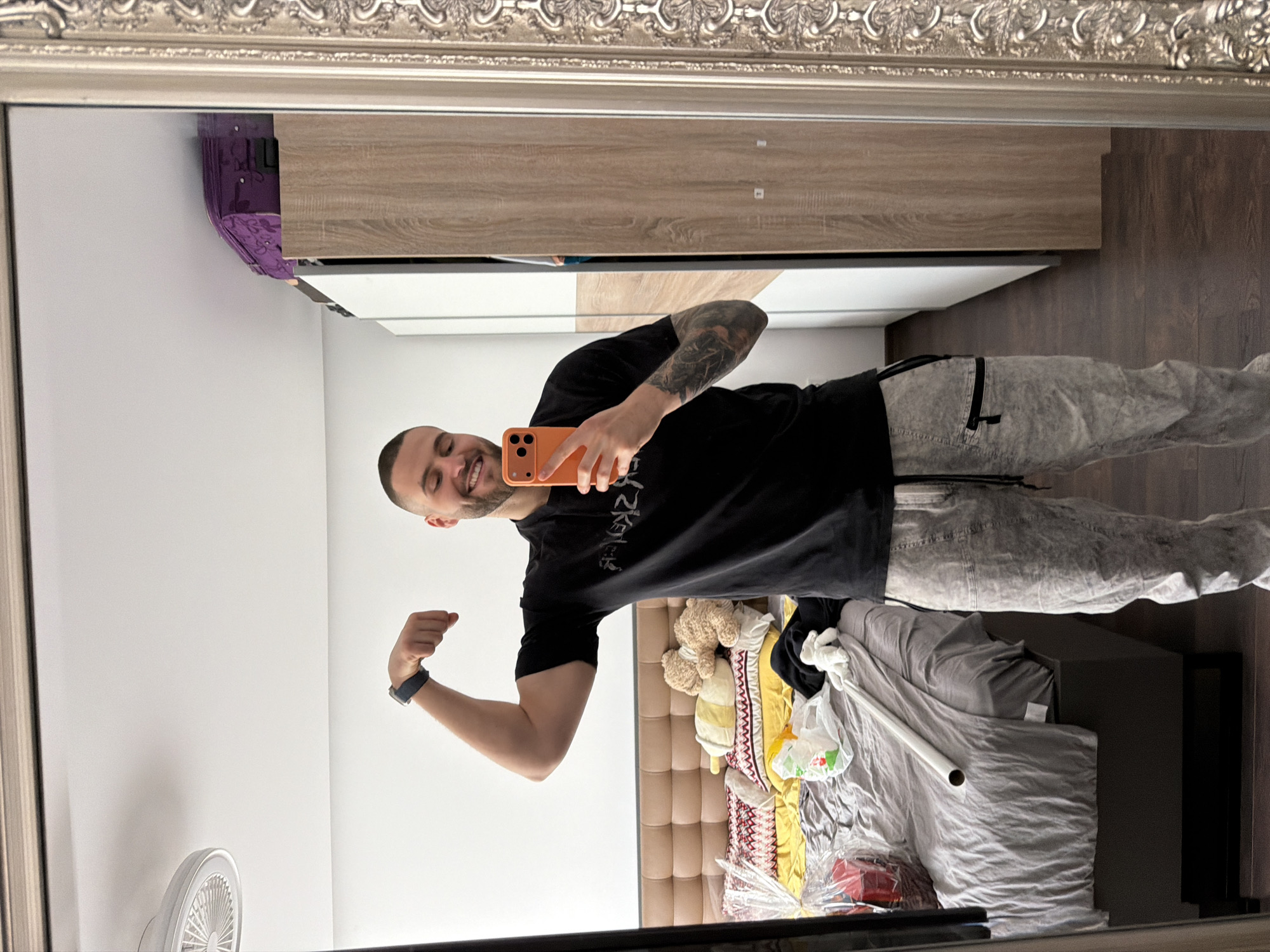A man is taking a selfie in a mirror inside an apartment. He is fair-skinned, has a shaved head, a short beard, and tattoos on his left arm. He is wearing a black t-shirt, grey pants, and a watch on his left wrist. He is holding an orange smartphone, presumably taking the photo. He is flexing his left bicep. The background includes a bed with scattered items, including a teddy bear, pillows, and a white tube. There is a closet on the wall. The location of this scene is Sofia, Bulgaria.
