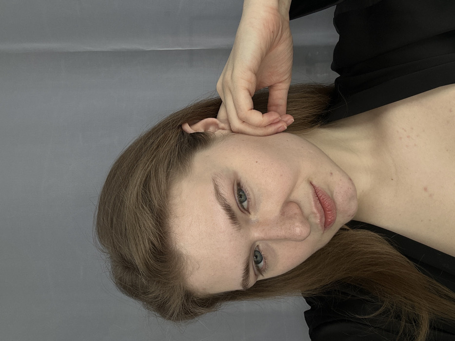 A fair-skinned woman with light brown hair poses in a sideways portrait. Her left hand is raised near her ear, fingers curled slightly. She has blue eyes and light eyebrows, with freckles visible on her face. She wears a dark garment, suggesting a robe or blouse. The background is a solid, neutral gray. The lighting is soft, highlighting her facial features and the texture of her hair.