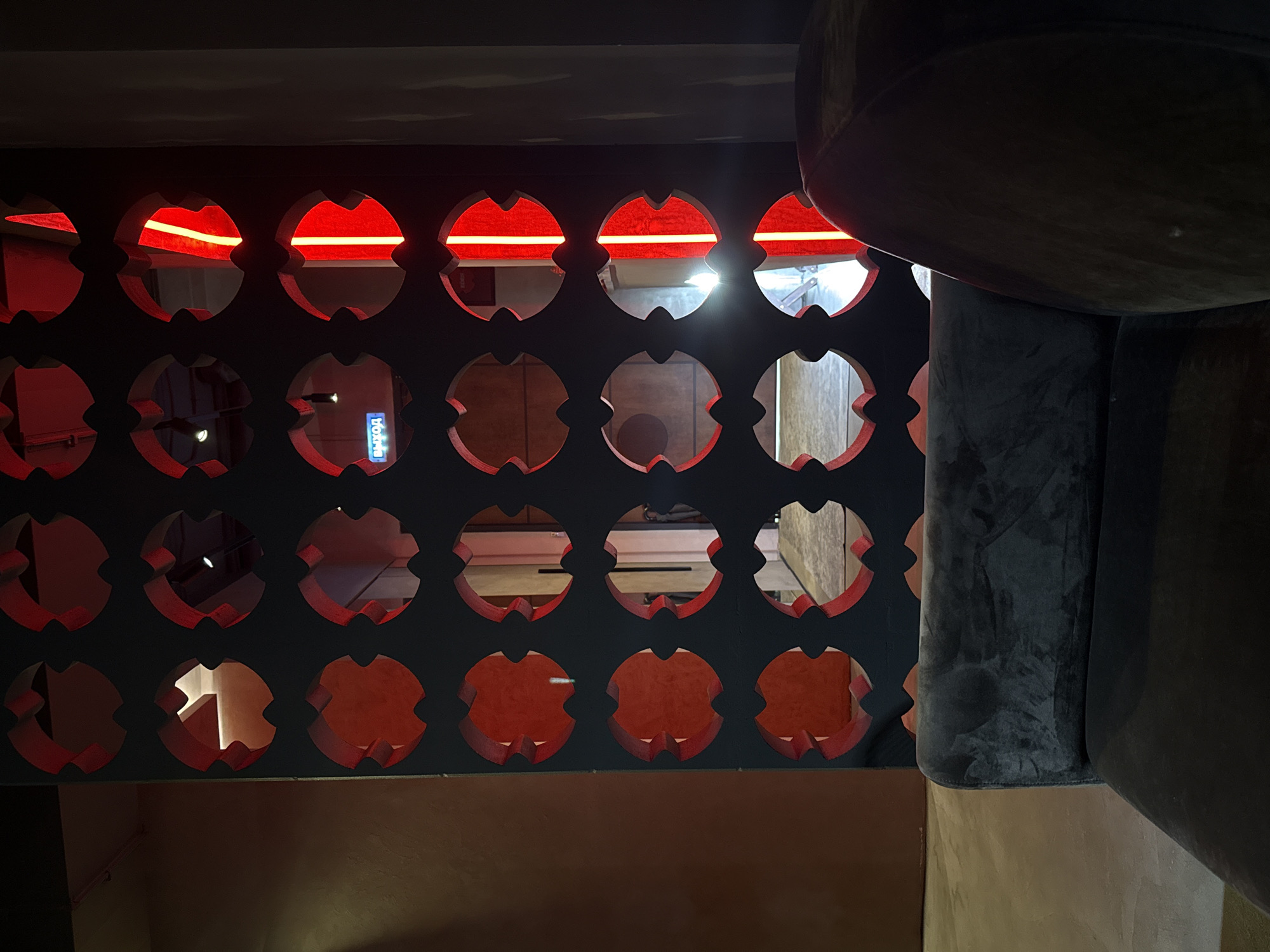 The image depicts a modern interior with a striking decorative wall panel featuring a repeating pattern of circular cutouts. Through these cutouts, glimpses of an adjacent space are visible, illuminated by red and white lights. One cutout clearly displays a neon blue sign that reads "ВЫХОД" (EXIT in Russian). In the foreground, a plush, dark-colored sofa or chair is partially visible, suggesting a seating area. The overall aesthetic is contemporary and somewhat moody, likely a commercial or hospitality space. This scene is located in Pushkino, Russia.