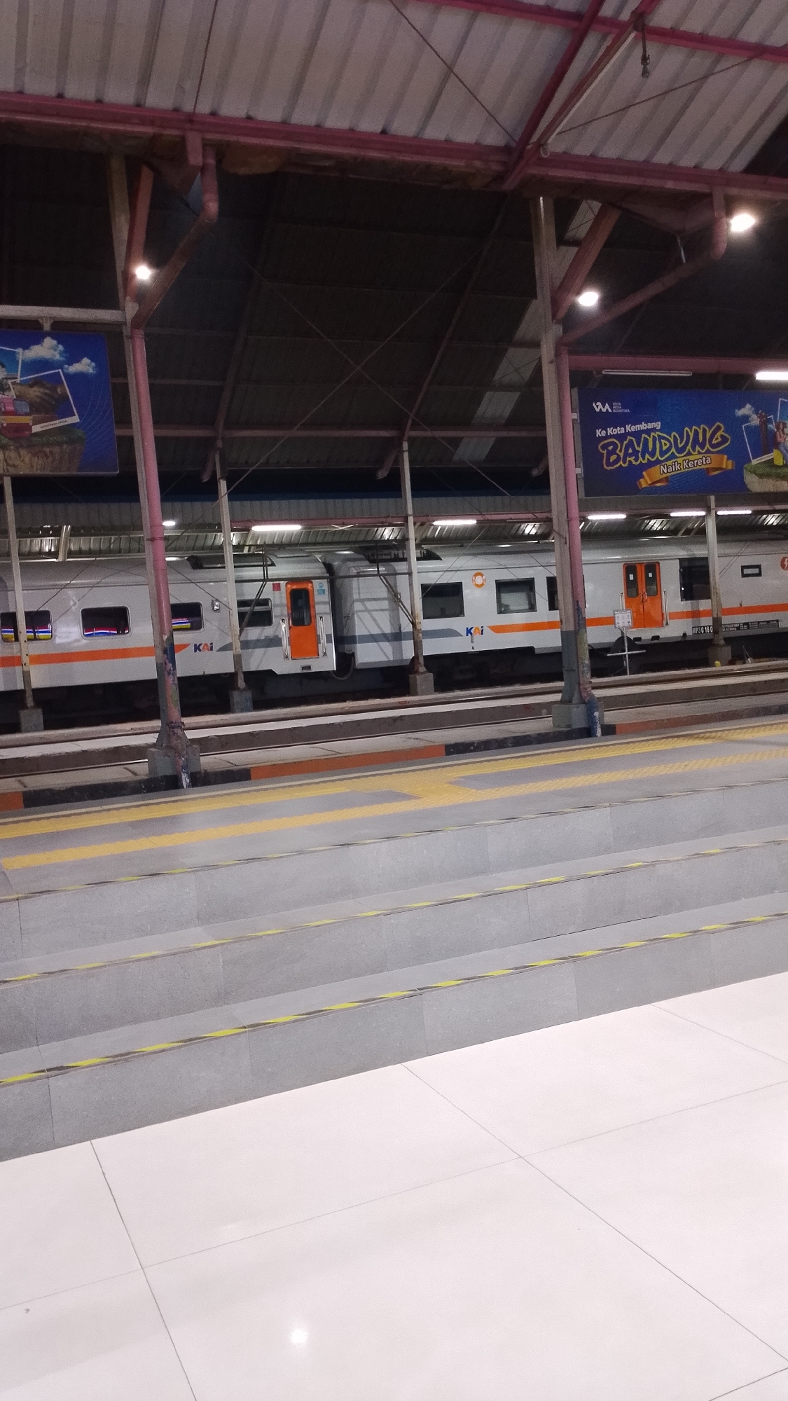 The image depicts a train station in Bandung, Indonesia. The foreground features a tiled floor and grey steps with yellow safety markings. Beyond the steps, a train is visible, stopped at a platform under a covered structure. The train cars are white and grey with orange doors and orange stripes. The word "KAI," the abbreviation for Kereta Api Indonesia (Indonesian Railways), is visible on the train. Above the train, advertising banners are displayed, one of which reads "Ke Kota Kembang BANDUNG Naik Kereta," which translates to "To the Flower City BANDUNG Take the Train." The station appears to be indoors or covered, with a metal roof supported by pillars. The lighting suggests it is either early morning or late evening.