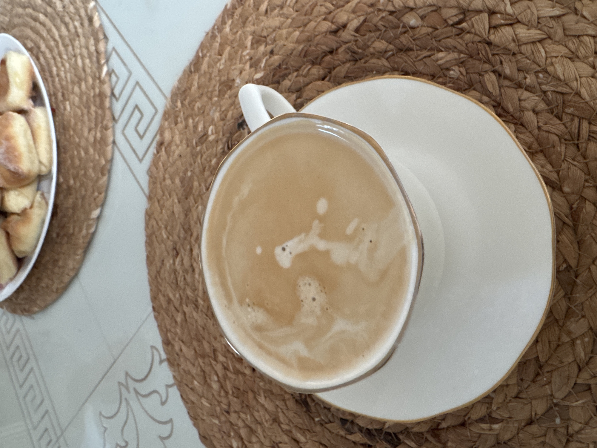 The image shows a cup of coffee on a white saucer with gold trim, sitting on a woven placemat. To the left is a plate of bread rolls, also on a woven placemat. The background consists of tiled flooring with a decorative pattern. There is no information to suggest that this image was taken in Izhevsk, Russia.