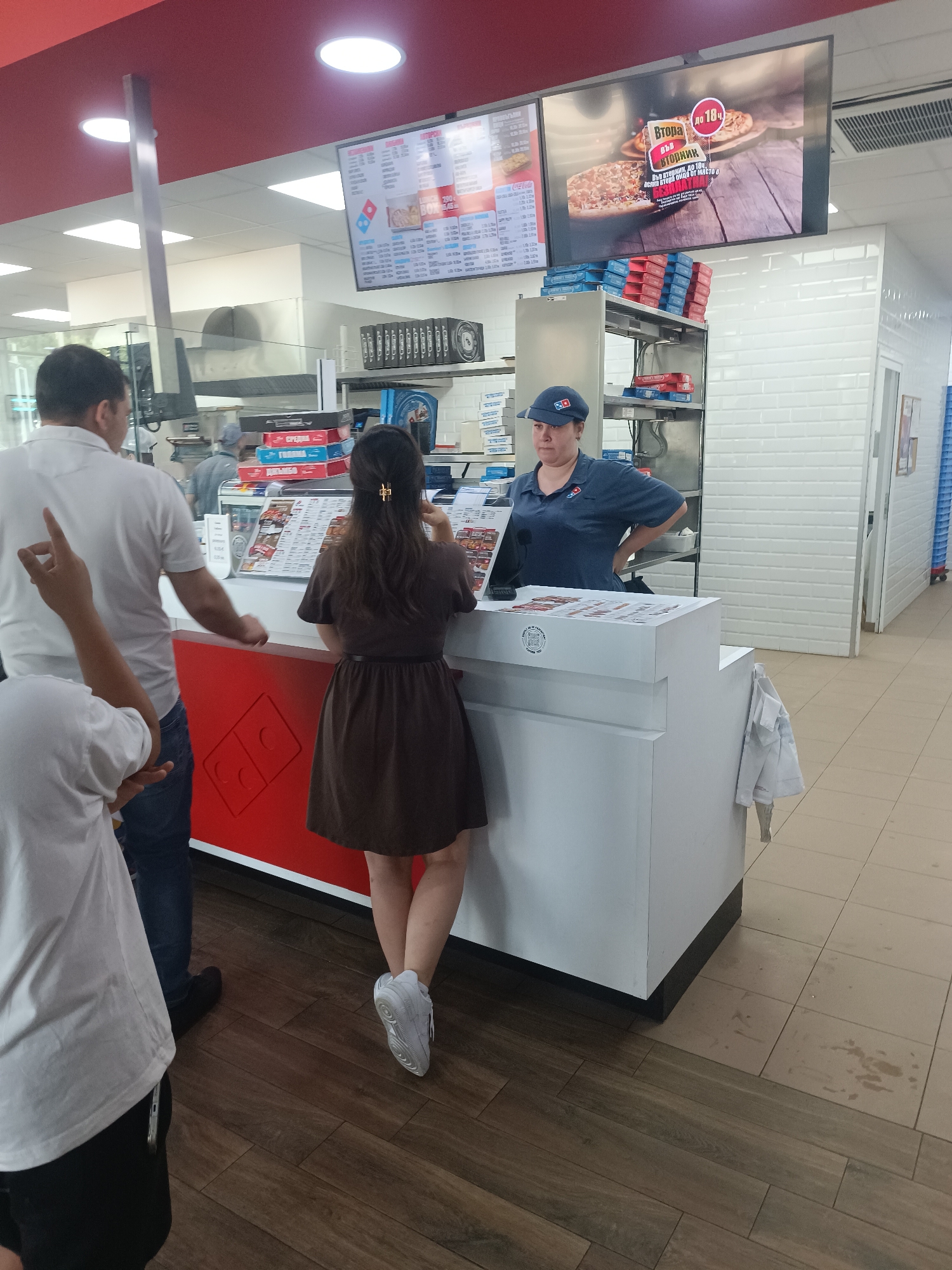 The image depicts the interior of a Domino's Pizza restaurant, likely located in Burgas, Bulgaria, as indicated by the Bulgarian text on the screen. The scene shows a counter area where customers are interacting with an employee.
At the counter, a woman in a blue Domino's uniform and cap is taking an order. She has her hands on her hips, facing the customers. In front of her, a woman in a dark brown dress with a gold hair clip is standing at the counter, appearing to be a customer. To the left of the counter, a man in a white t-shirt and jeans is standing, and his left arm is raised with his index finger pointed. Another person, partially visible from the chest down, is standing to the far left, wearing a white t-shirt and black shorts, with their left hand raised as if pointing or gesturing.
Behind the counter, shelves are stocked with pizza boxes. A large screen is mounted on the wall above the counter, displaying an advertisement for a pizza promotion with Bulgarian text. The counter itself is white with a red section featuring the Domino's logo. The floor is a combination of wood-patterned tiles and light-colored ceramic tiles. The overall environment suggests a busy fast-food establishment.