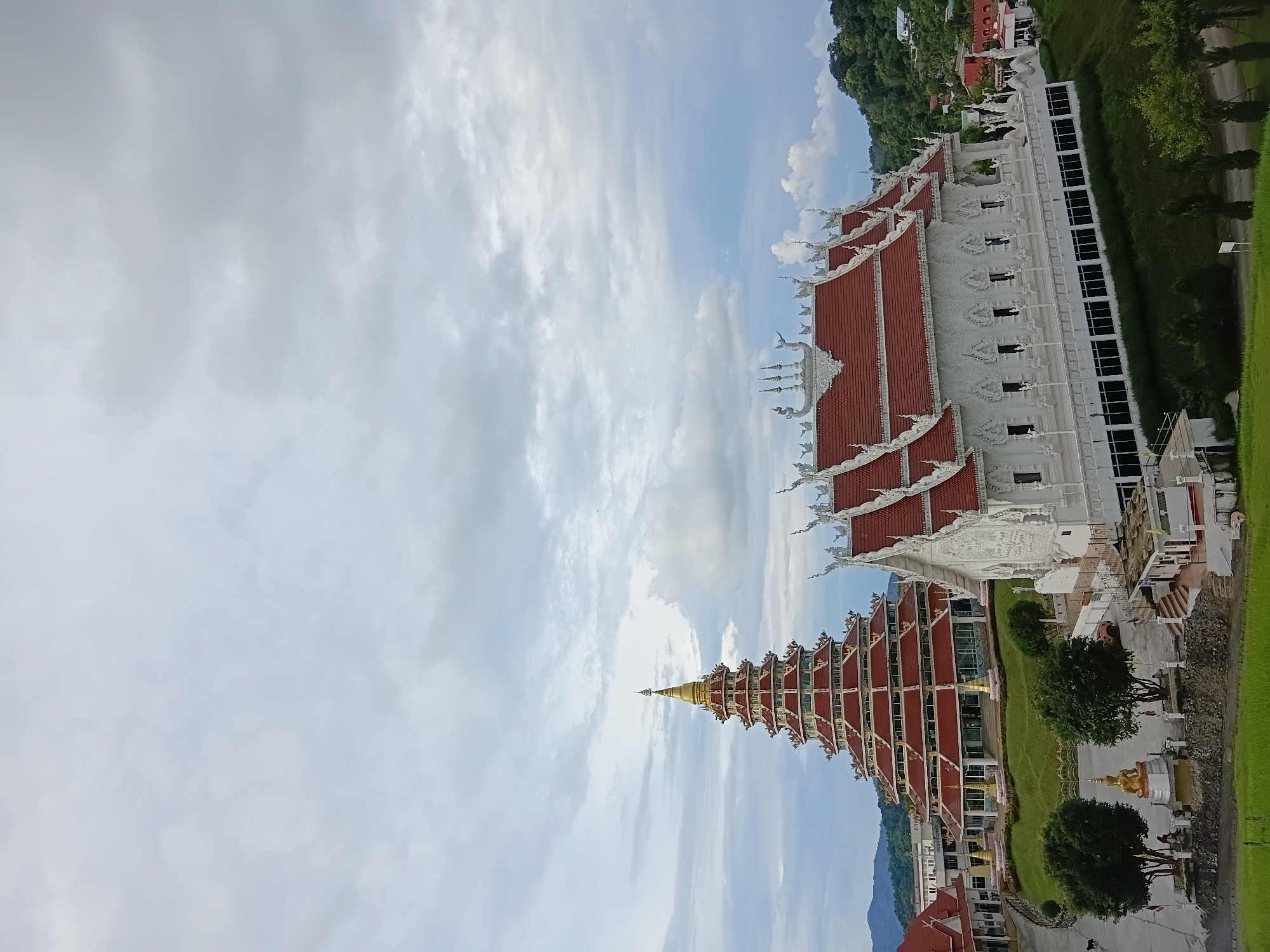 The image depicts a temple complex nestled in a hilly landscape under a cloudy sky. In the foreground, a white temple with intricate carvings and a red tiled roof dominates the scene. To the left, a multi-tiered, golden-capped stupa with red and gold accents rises majestically. The temple grounds are green and landscaped, with trees and pathways. A statue of a golden Buddha is visible in front of the stupa. In the background, more buildings of the temple complex and lush green hills are seen. The sky is filled with dramatic clouds, suggesting a potentially overcast or stormy day. The overall impression is one of serene grandeur and architectural splendor. The location is identified as Wat Huay Pla Kang in Ban Lao Phatthana, Mueang, Chiang Rai, Thailand.