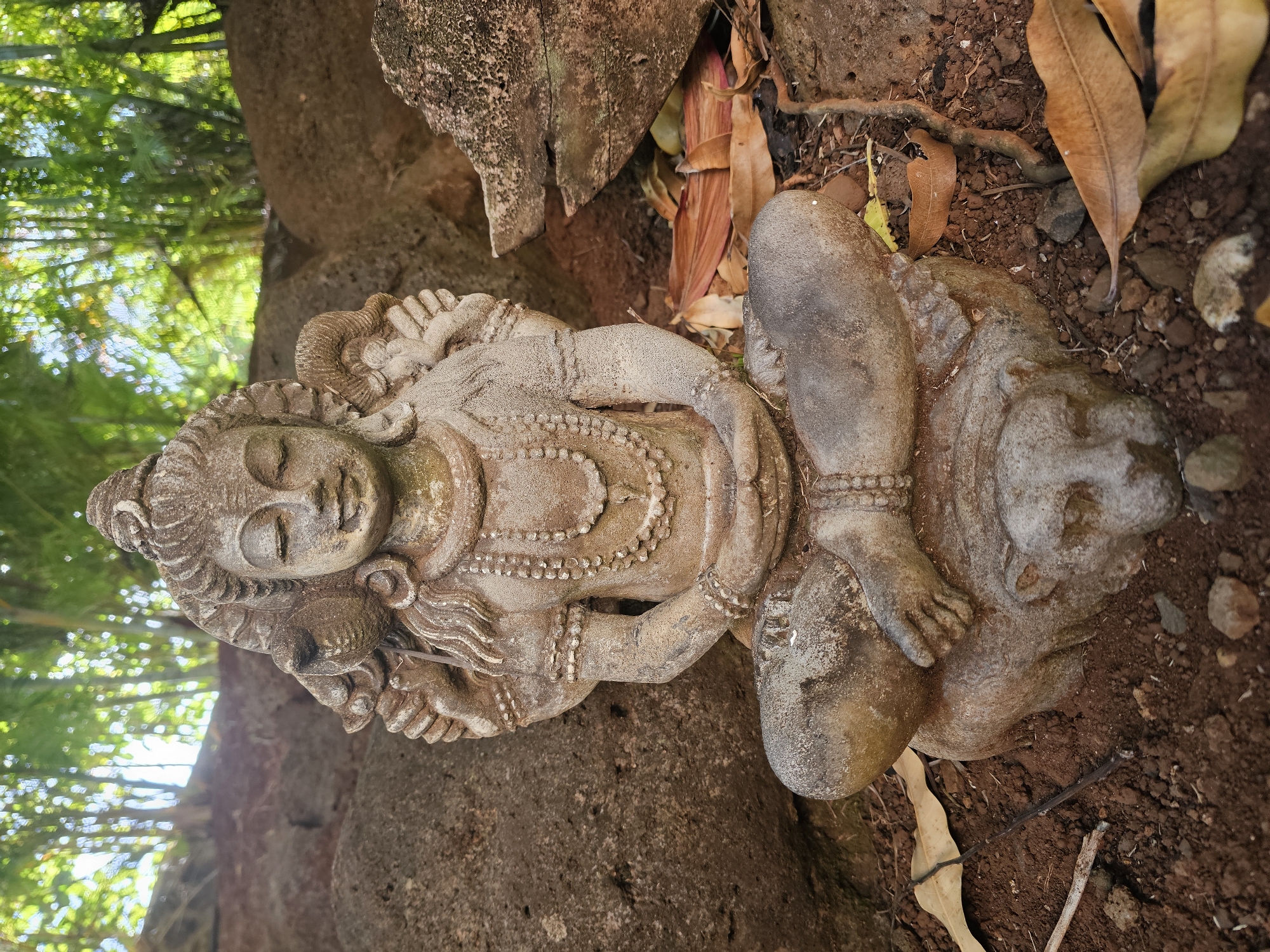 A stone statue of a multi-armed deity, possibly Shiva, is seated in a meditative pose on a stone base that appears to be carved into the form of a reclining animal. The statue is positioned outdoors, surrounded by rough-hewn rocks and loose soil. Dry leaves are scattered on the ground around the statue, and in the background, a dense growth of green foliage and trees is visible, with sunlight filtering through the leaves. The statue exhibits a weathered appearance, suggesting it has been exposed to the elements. The setting suggests a natural or garden environment.