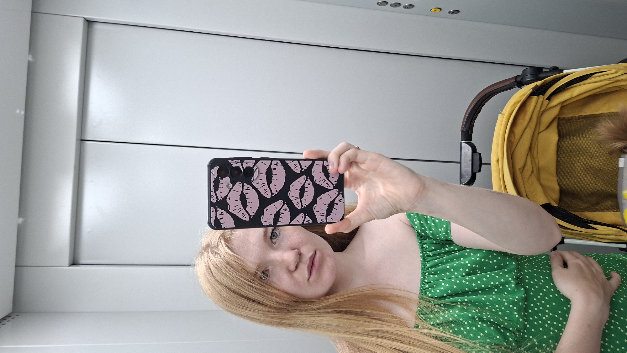 A young woman with long blonde hair and fair skin is taking a selfie in an elevator. She is wearing a green off-the-shoulder dress with white polka dots. She is holding up a smartphone with a black case featuring a pattern of pink lips. To her right, a yellow baby stroller is visible. The elevator walls are grey and metallic. The image appears to be taken in an urban environment, possibly in Chaikovsky, Russia, given the mention of the city.