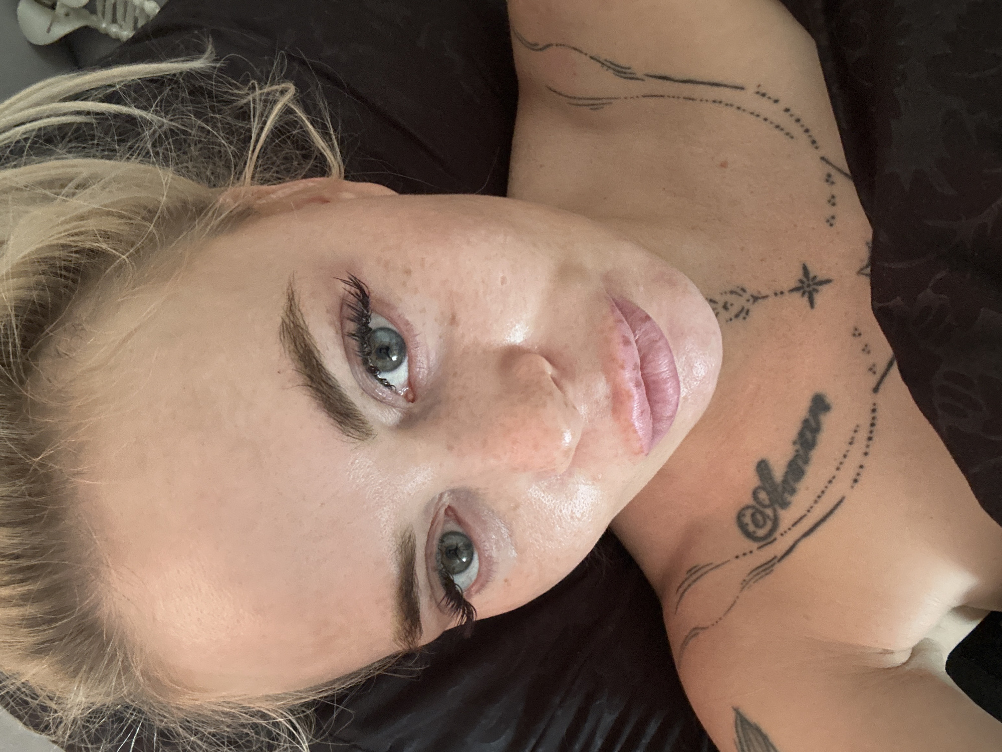 A close-up, overhead shot shows a fair-skinned woman with blonde hair pulled back. She has blue-green eyes, light brown eyebrows, and is wearing false eyelashes. Her skin is visible with freckles and some redness around her nose and cheeks. Her lips appear plump and are lightly colored. She is wearing makeup, including eyeshadow and possibly blush. There are tattoos visible on her chest and shoulder area. One tattoo on her collarbone reads "Amor" in cursive, and another on her chest depicts a star and circular patterns. She is lying down on a dark, possibly satin surface, with a glimpse of a skeletal decoration in the upper right background. The lighting suggests an indoor environment.