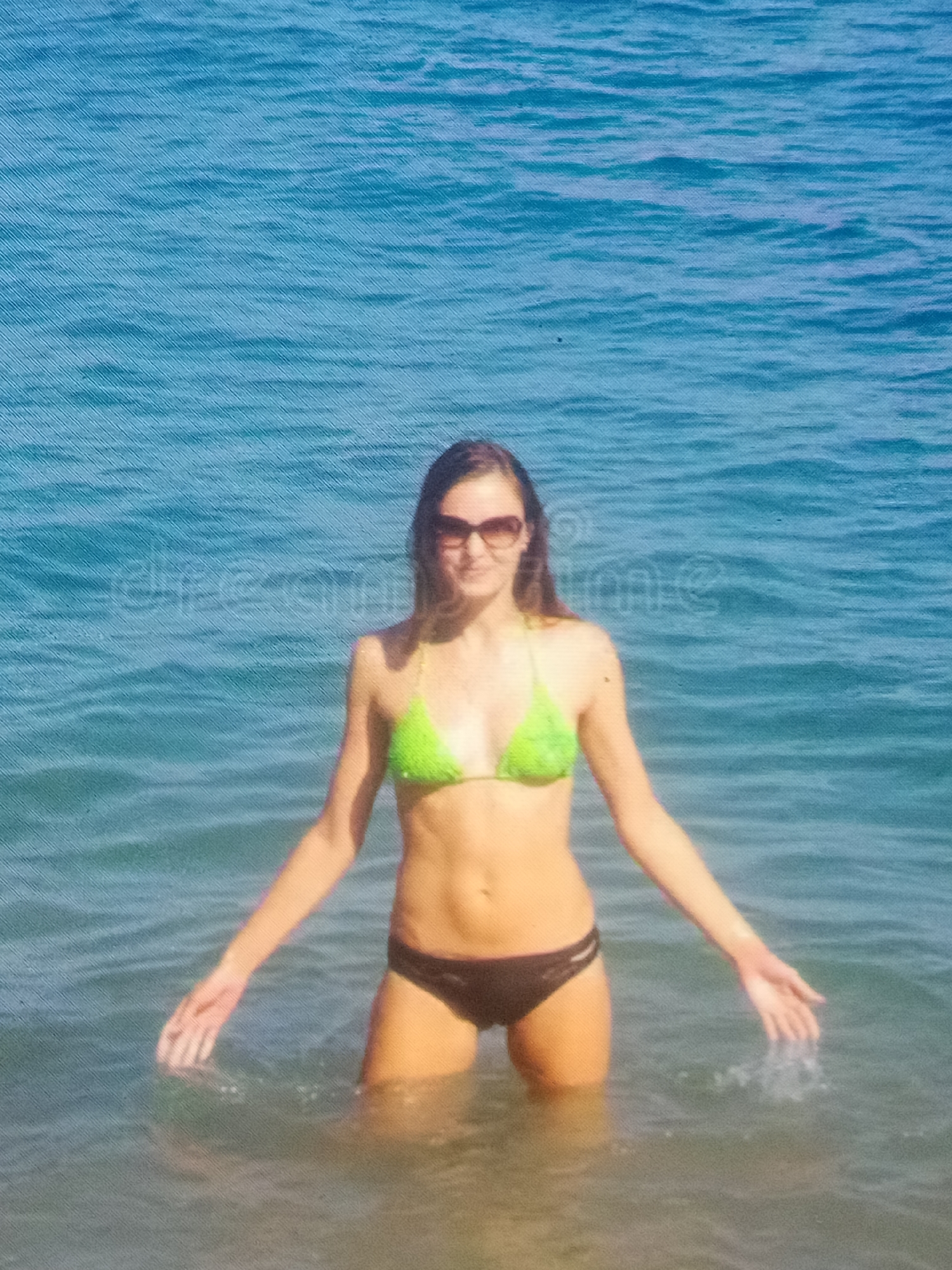 A forensic examination of the image reveals a lone female subject standing waist-deep in a body of blue water, consistent with a large lake or ocean, located in Unknown, Unknown. The subject, a young adult female with long, dark brown hair, is wearing a lime green halter-neck bikini top and dark (likely black or dark brown) bikini bottoms. She is also wearing dark-lensed sunglasses. Her arms are extended horizontally from her sides, with her hands open and palms slightly downwards, just above the water surface, creating small disturbances on the water. The water exhibits a ripple pattern, and the color transitions from a lighter blue in the foreground to a deeper blue in the background. A faint, semi-transparent "dreamstime" watermark is visible horizontally across the mid-upper portion of the image. The image itself shows a textured pattern, possibly indicative of a scanned print or a photograph of a screen.