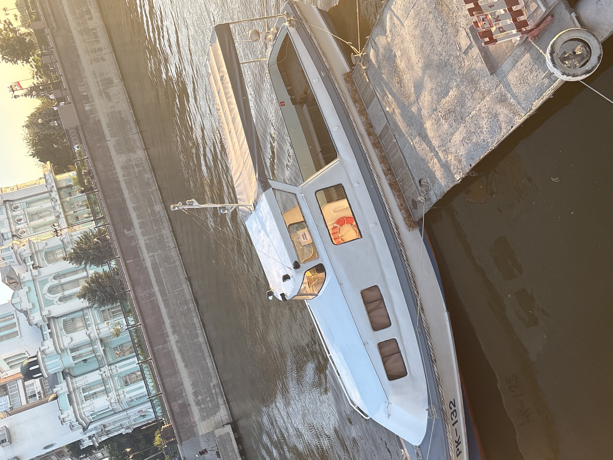 The image shows a white passenger boat docked on a canal in Astrakhan, Russia. The boat is modern in design with large windows and a covered upper deck. It appears to be at rest, possibly awaiting passengers or undergoing maintenance. The scene is bathed in the warm light of either sunrise or sunset, casting long shadows and creating a serene atmosphere.
In the background, an ornate, pale blue and white building with classical architectural elements lines the canal. Trees and lampposts are visible along the embankment, suggesting a public space. The water in the canal is calm, reflecting the sky and the surrounding structures. The time of day is likely late afternoon or early morning due to the low angle of the sun and the warm, golden light.
Visible text on the boat includes "PK-132" on the side and what appears to be "PK-105" reflected in the water. There are also some smaller red signs inside the boat's cabin that are too small to read clearly. The overall impression is one of a peaceful urban waterway scene, typical of a European city during a pleasant time of day. There are no visible people in the image, suggesting the boat is either empty or the activity is not centered around immediate passenger boarding.