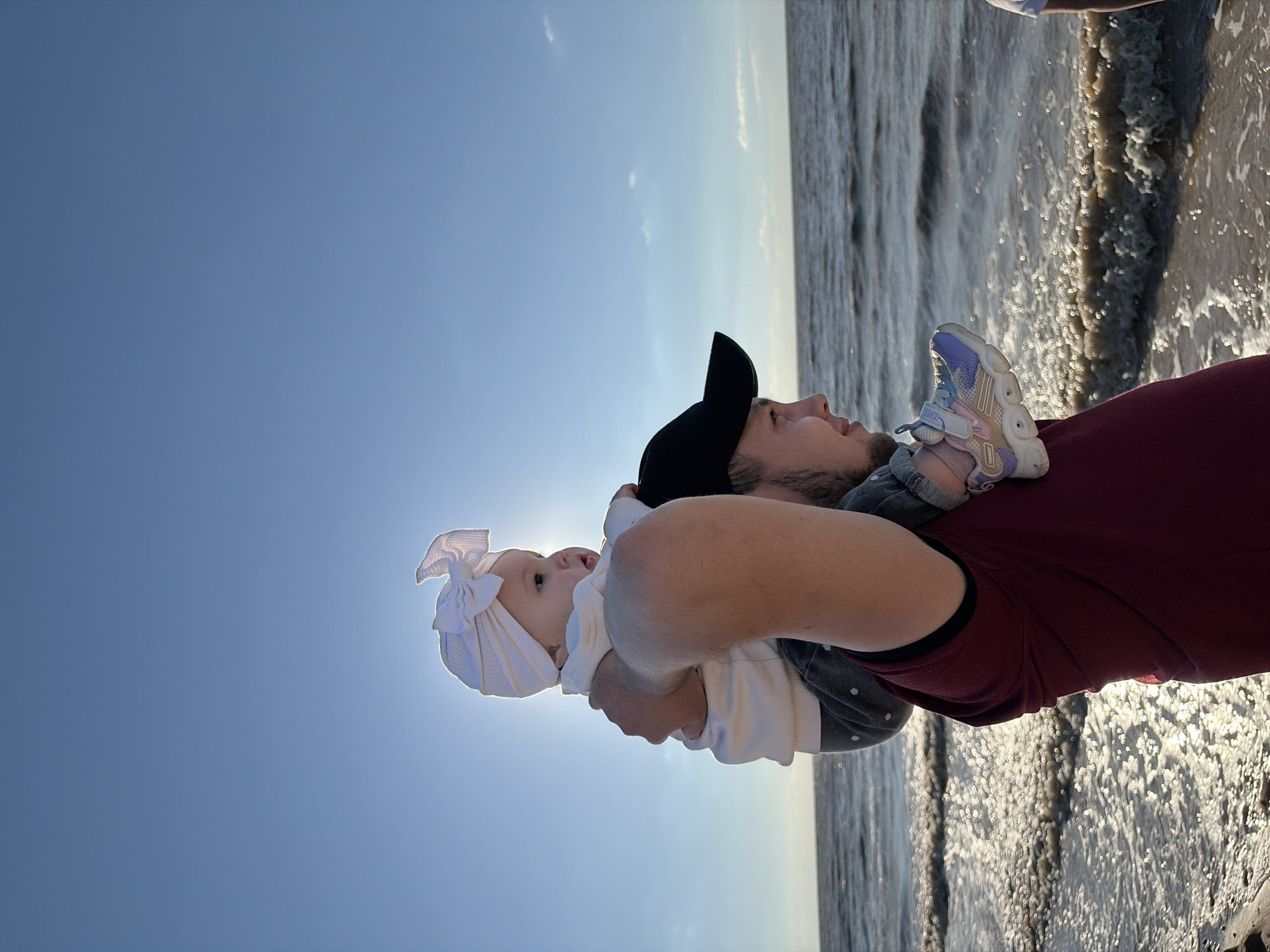 In Port-Katon, Russia, a man wearing a maroon t-shirt and a black baseball cap carries a baby on his shoulders. The baby, adorned with a white turban, gazes towards the ocean. The sun is visible behind the baby, casting a bright glow. The water's surface reflects the sunlight, creating a shimmering effect. The scene suggests a family enjoying a day at the beach.