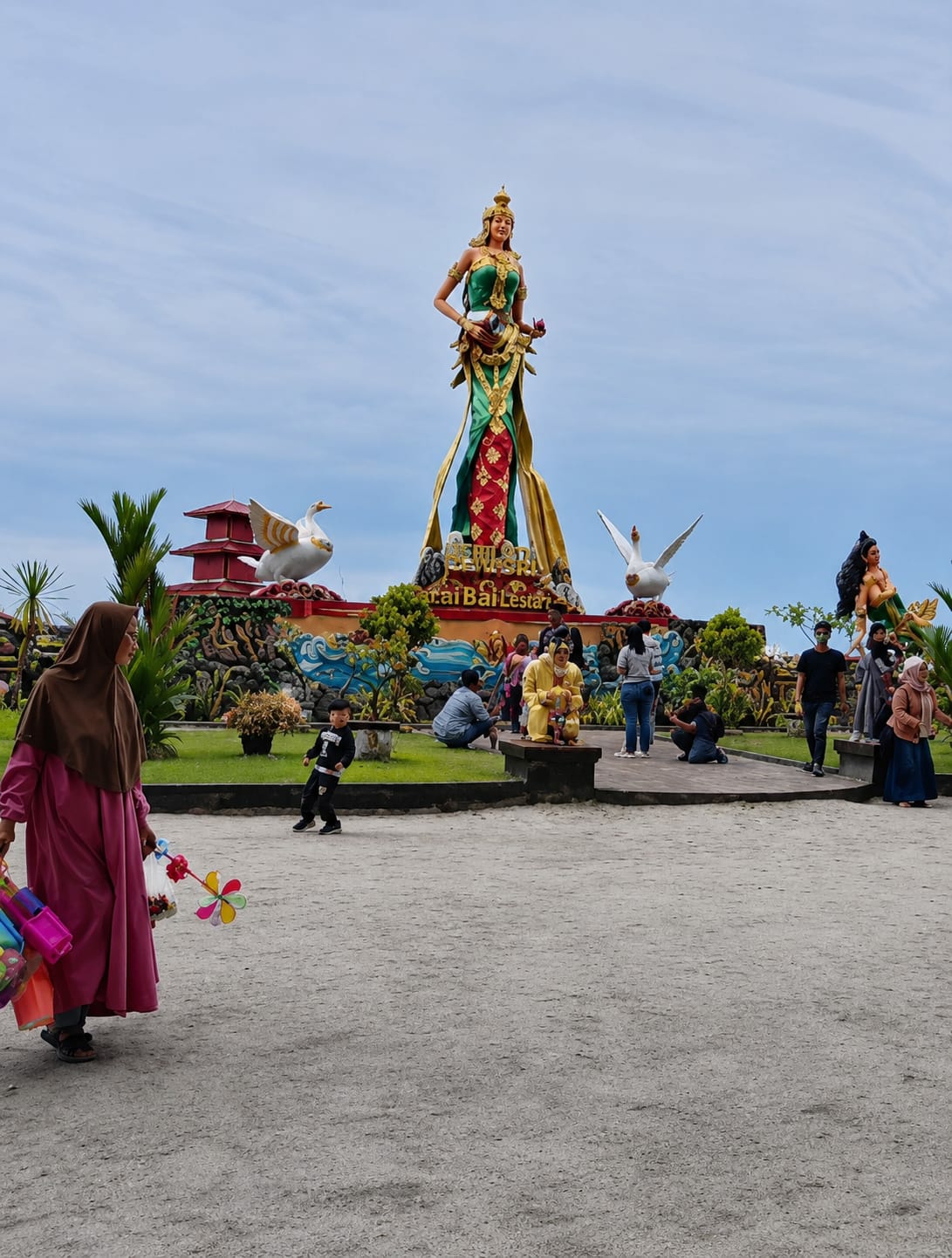 The image depicts a sunny day at a public square in Kota Pari, Indonesia, featuring a large, vibrant statue of a woman in traditional attire. The statue is adorned with green and red fabric, with gold accents, and is positioned on a raised platform. Flanking the central statue are two large, white swan statues with yellow beaks and wings, one on either side. In the background, a red and white tiered building with white ornamental birds is visible. A group of people are gathered around the statues, some seated, some standing, and some taking pictures. In the foreground, a woman in a pink hijab and a long pink dress is walking from left to right, carrying a collection of colorful pinwheels and other toys. A young boy in a black shirt and pants is running in the middle ground. The ground is a light-colored gravel path. The sky is partly cloudy with a clear blue expanse.