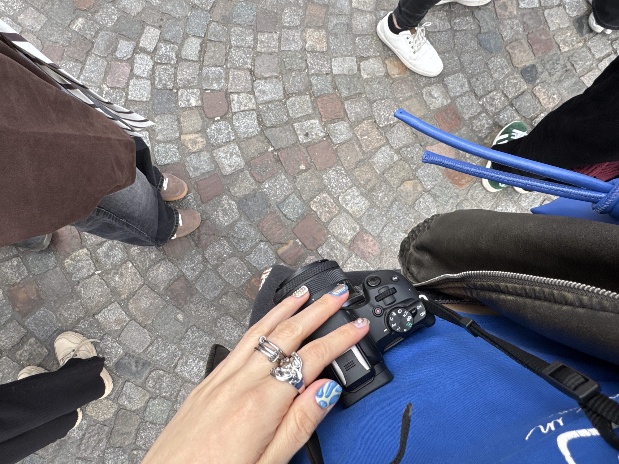 Captured from an elevated perspective, the photograph presents a view of a cobblestone street, presumably located in a European urban setting, possibly Paris, France, based on the architectural style of the paving stones and anecdotal context.
Four individuals are partially visible in the frame. One wears brown outerwear, dark jeans, and brown shoes. Another is wearing a white athletic shoe. A third individual is shown wearing black pants and green and white shoes.
The focal point of the image is a person holding a black camera with an attached lens and a blue handle. The person is wearing a blue shirt. The hand holding the camera is adorned with several silver rings and manicured nails with blue designs. The camera rests on the person's chest, which is covered with a gray bag and a blue top.
The ground is covered with cobblestones of varying colors and shapes, suggesting an older part of the city.