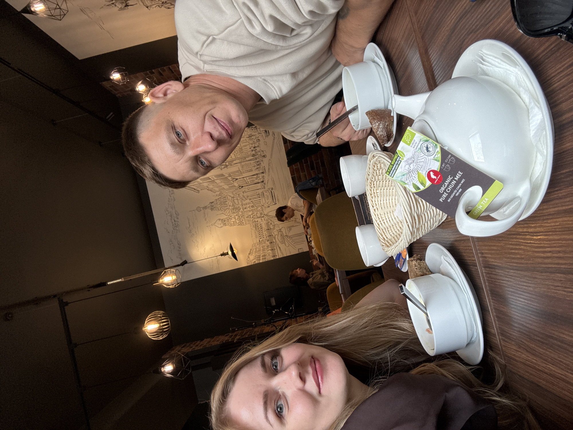 Two individuals, a man and a woman, are seated at a wooden table in a café, presumably in Saint Petersburg, Russia. The man is fair-skinned with short hair and a light-colored shirt. The woman has long blonde hair and is wearing a dark-colored top. Tea service items, including cups, saucers, a teapot, and a tea bag wrapper for "Organic Pure Chun Mee Green Tea," are arranged on the table. A slice of bread sits nearby. The café's interior features exposed brick, decorative light fixtures, and a mural depicting the city's architecture. Other patrons are seated at tables in the background.