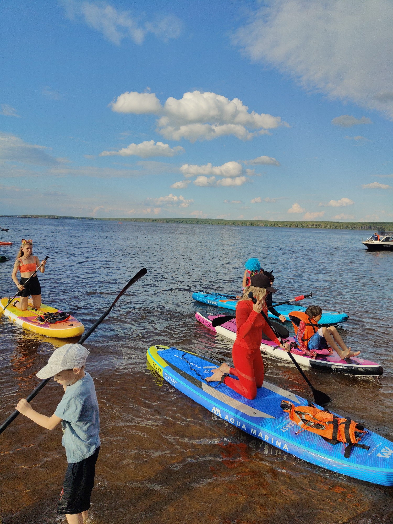 This media file depicts a group of people paddleboarding on a body of water under a bright, partly cloudy sky. The scene appears to be a recreational outing on a lake or calm sea. Several individuals are visible on paddleboards, some kneeling and others sitting. One person, wearing a bright red wetsuit, is kneeling on a blue paddleboard. A child in an orange life vest is sitting on a pink paddleboard. Another young person in a blue t-shirt and black shorts is standing in the shallow water, holding a paddle. In the background, a boat is visible on the water, and a tree line forms the horizon.
The weather seems to be pleasant, with a blue sky dotted with white clouds, suggesting a sunny day. The water is a deep blue, and the reflections on its surface indicate calm conditions. The time of day appears to be afternoon, given the brightness of the sunlight.
Visible text on the paddleboards includes "ICE FOGL", "SPORTS II", "BEAST", "BEAST 10'6\"x32\"", and "Aqua Marina". The overall mood of the image is one of leisure and outdoor activity.
