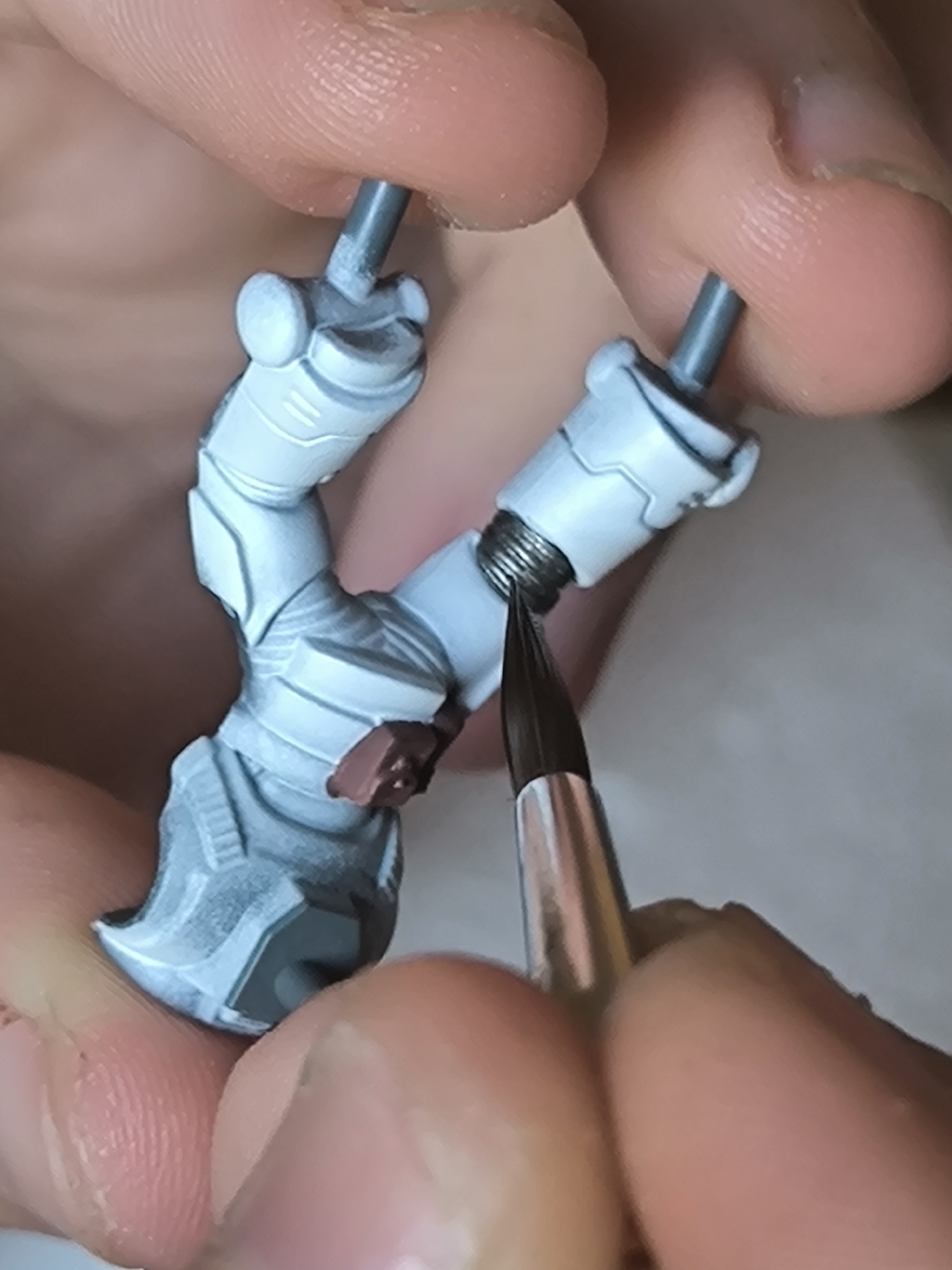 Close-up of a person's hands holding and painting a miniature model figure. The figure appears to be a futuristic or sci-fi soldier with articulated joints and armor plating. The armor is painted in a pale gray or white color with some details in darker gray. One joint has a silver coil detail. A paint brush is being used to apply paint to the figure. The background is blurry.