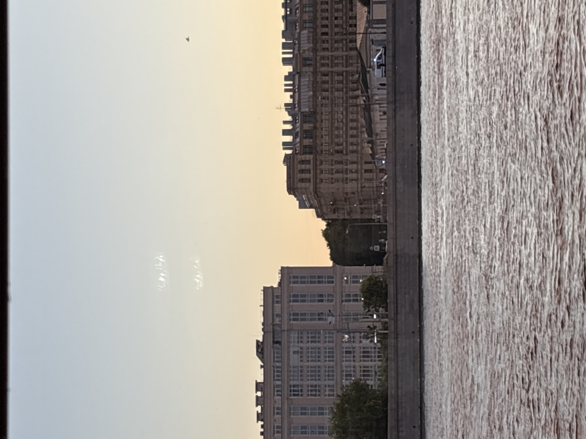 The image shows a waterfront view of two large buildings in an urban setting. The building on the left is modern, with a white facade and numerous windows. The building on the right is older, with a more ornate, tan facade and many chimneys along the roofline. Between the two buildings, there is a gap with trees and a darker, smaller building. In the foreground, there is a wide body of water with visible ripples, suggesting movement. A stone embankment separates the water from the buildings. The sky is a pale gradient of blue and yellow, indicating either early morning or late evening light. There are some blurry streaks or smudges in the sky, possibly from dust or reflections. A small dark shape, potentially a bird or an aircraft, is visible in the upper right portion of the sky. A white vehicle, possibly a police car due to its lights, is parked near the building on the right. There are no people clearly visible in the image.