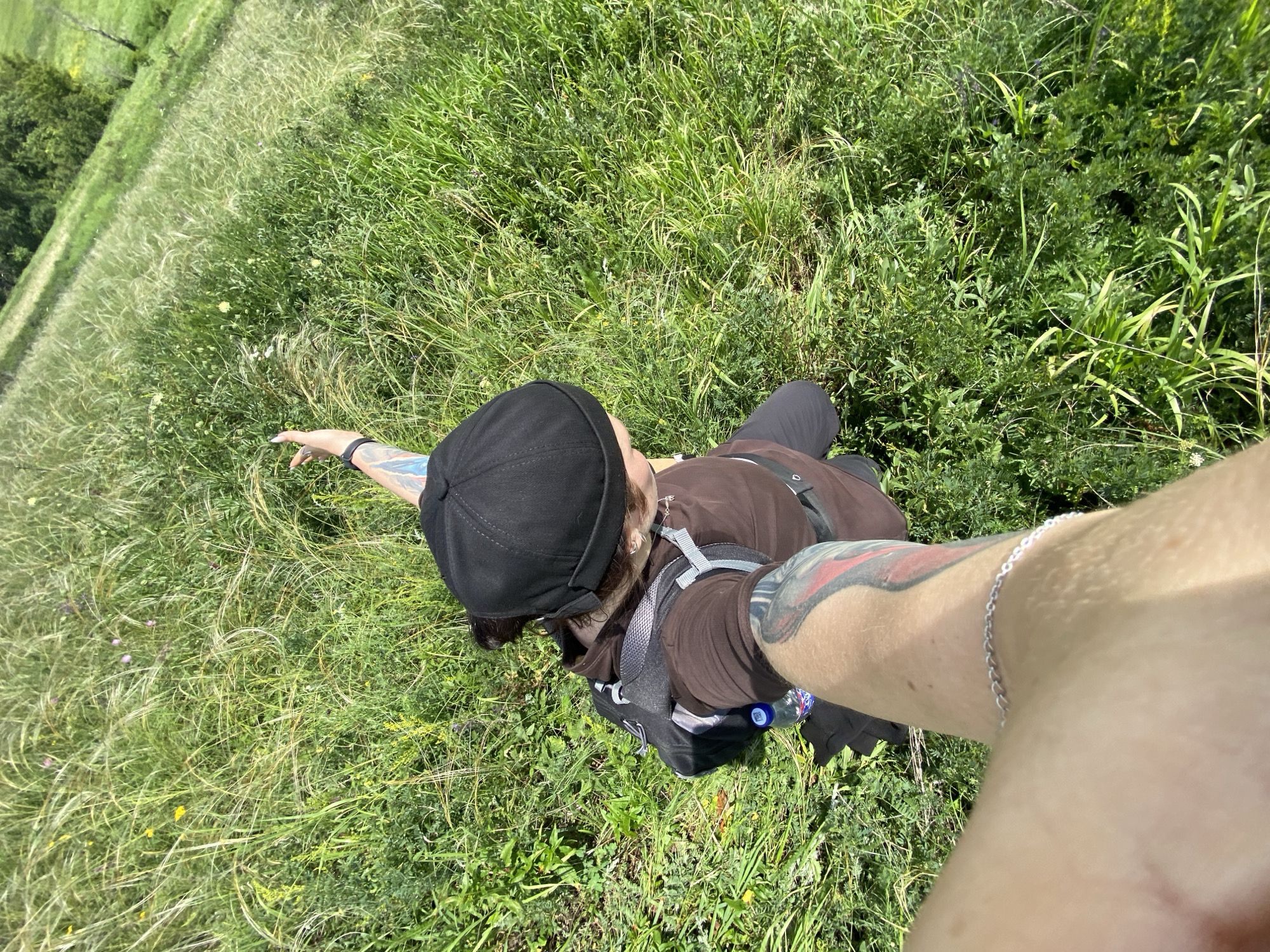 An overhead, slightly angled, point-of-view shot shows a person walking on a grassy hillside. The person is wearing a brown long-sleeved shirt, dark pants, and a black beanie. A backpack is on the person's back, and a water bottle is visible attached to the backpack strap. The person's right arm, extended towards the camera, is tattooed and adorned with a silver bracelet. The environment is a lush green hillside covered in tall grass and some scattered wildflowers. The background shows more hillside and a patch of trees. This scene is consistent with the landscape found in the city of Bogoslovka, Russia, which is known for its natural beauty and rolling hills.