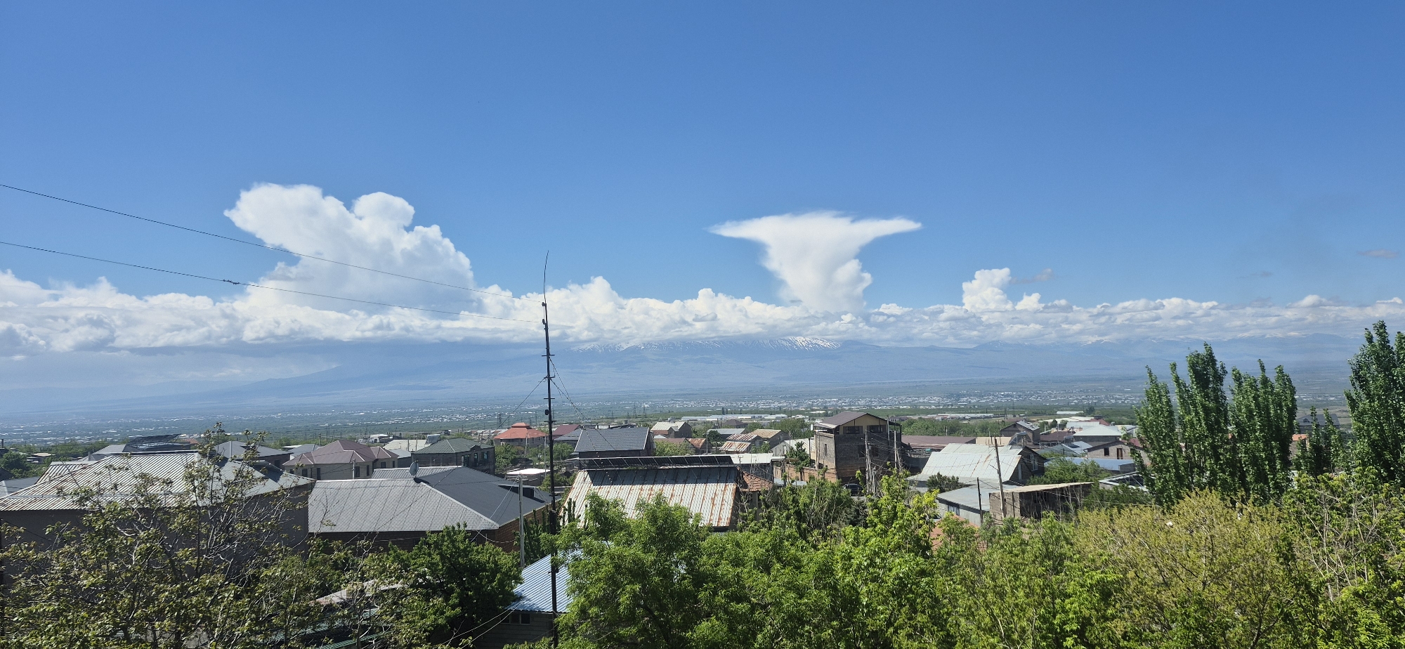 The image depicts a daytime view of a town located in the city of Unknown, Unknown, with a clear blue sky partially covered by cumulus clouds. A large cumulonimbus incus cloud is seen in the distance, rising above a mountain range with visible snow-capped peaks. The town features a mix of residential buildings with various roof types, surrounded by green trees and foliage. A tall telecommunication antenna is centrally positioned in the middle ground. Power lines stretch across the upper left portion of the frame.
