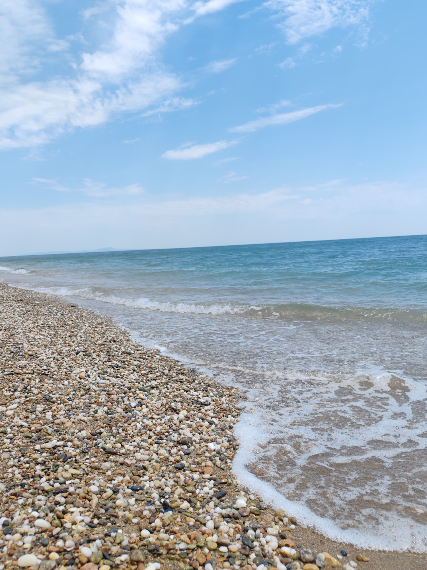 A pebble beach with the Mediterranean Sea and a light blue sky with wispy clouds. The pebbles are smooth and rounded, in various shades of brown, gray, white, and black. The sea is a clear blue-green with gentle waves lapping at the shore. The sky is a soft blue with scattered white clouds. The image appears to have been taken on a sunny day. The location is the beach in Makri, Greece.
