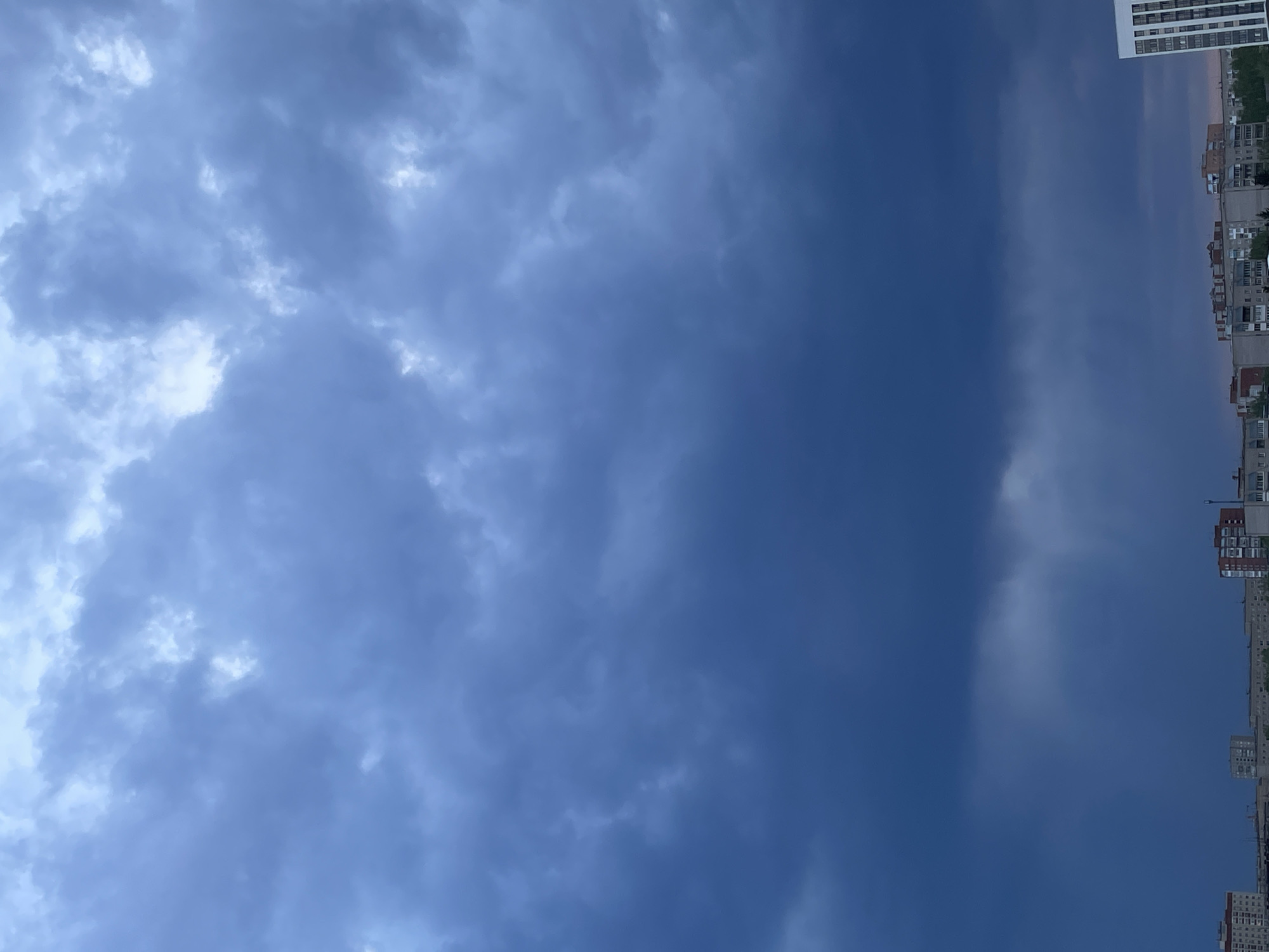 The image presents an upward view of a sky filled with clouds, appearing overcast and stormy. The clouds range from light grey to dark blue, creating a dramatic contrast. Along the right edge of the frame, the tops of buildings are visible, suggesting an urban environment. The buildings are a mix of residential and commercial structures, typical of cityscapes. The specific location appears to be in Izhevsk, Russia, based on architectural styles. The lighting indicates it may be late afternoon or early evening on a cloudy day. No people are visible in the frame.