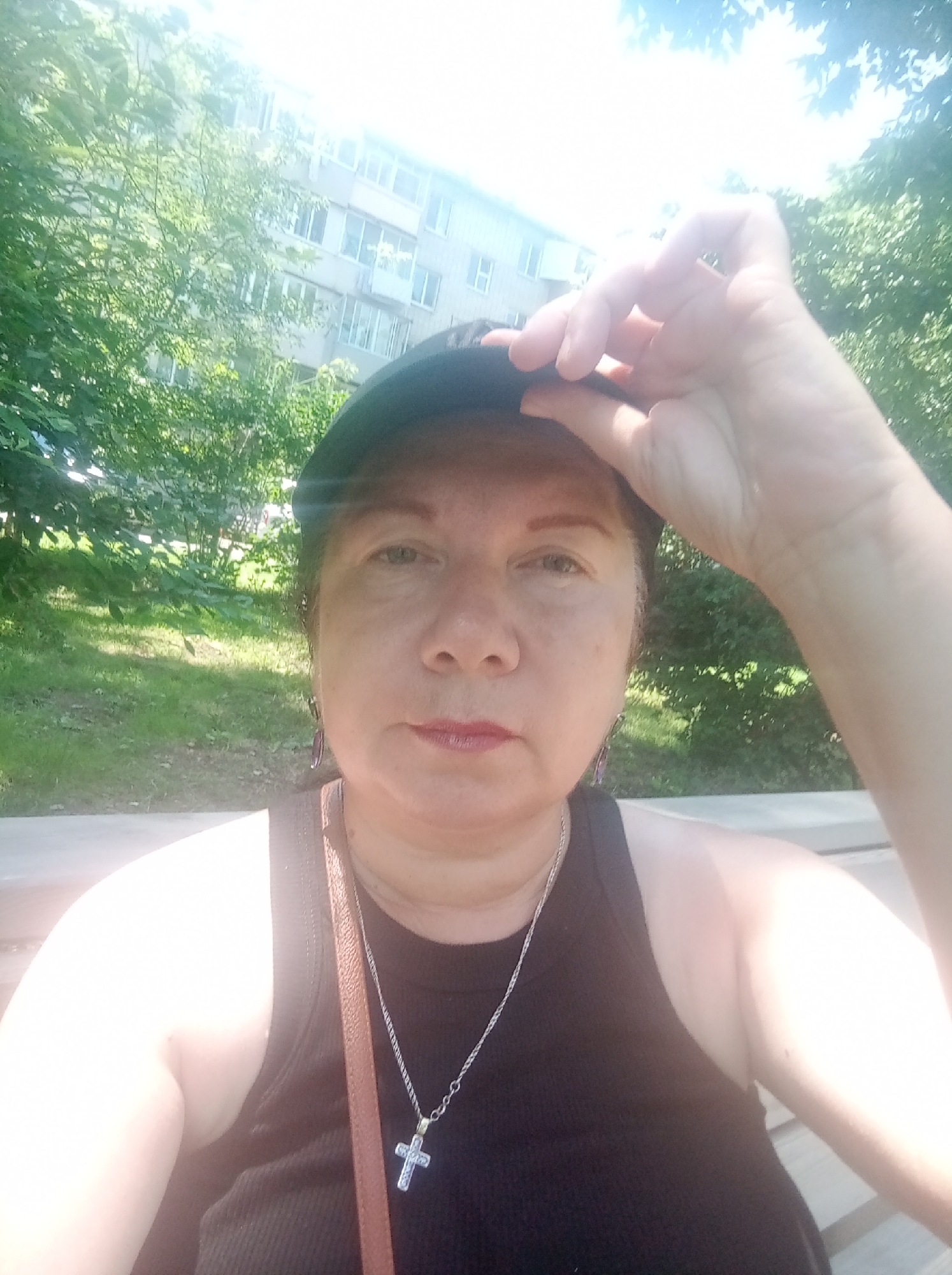A woman wearing a black tank top and a black baseball cap is taking a selfie outdoors. She has her right hand on the brim of her cap, and is wearing a silver necklace with a cross pendant. A brown purse strap is visible across her shoulder. She is sitting on a wooden bench, with trees and a building in the background. The building appears to be a multi-story apartment complex. The image was taken in the city of Artyom, Russia.