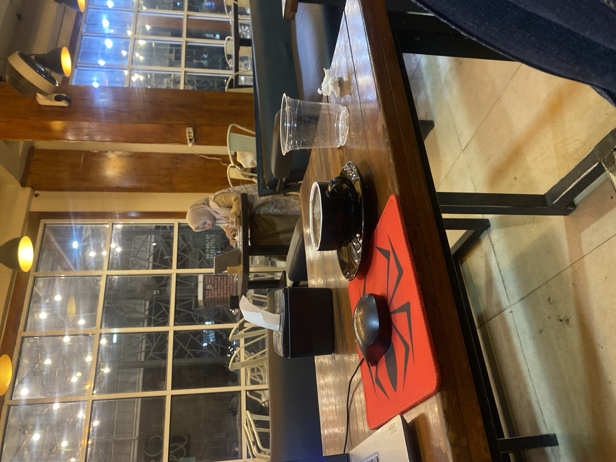 A woman wearing a hijab is seated at a table in a dimly lit cafe, illuminated by overhead lights and the reflections from a large window. She is looking down at a laptop. In the foreground, a wooden table holds a cup of coffee, an empty plastic cup, a tissue dispenser, a computer mouse on a red mousepad, and a laptop. The cafe appears to have a modern, industrial aesthetic with exposed wooden beams and dark furniture. The scene is set in Lhokseumawe, Indonesia.