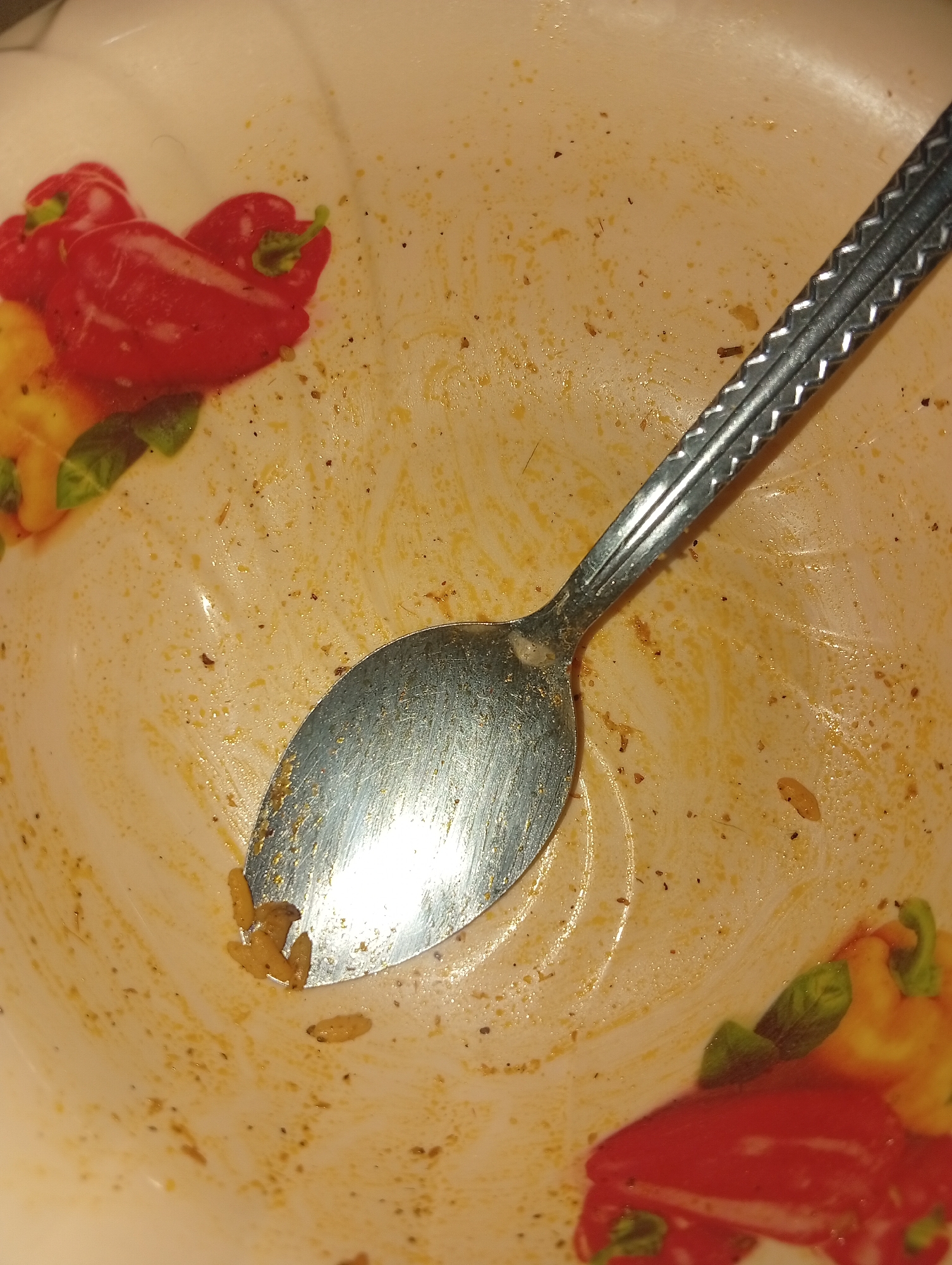 The image displays a close-up view of a dirty white ceramic bowl with a pattern of red and yellow bell peppers and green leaves. A silver metal spoon rests in the bowl, which contains remnants of a yellowish-orange food and some scattered dark specks that appear to be spices or small pieces of food. Several small, light-colored, curved objects, resembling pieces of pasta or grains, are also visible in the bowl, particularly near the spoon. The surface of the bowl is wet and has streaks of the food residue. The lighting is warm and somewhat dim, creating reflections on the spoon and the wet surfaces of the bowl. The scene is domestic, likely a kitchen or dining area, but the specific location in Ukukwa, Nigeria, cannot be determined from the image itself. There are no people or animals present in the image.