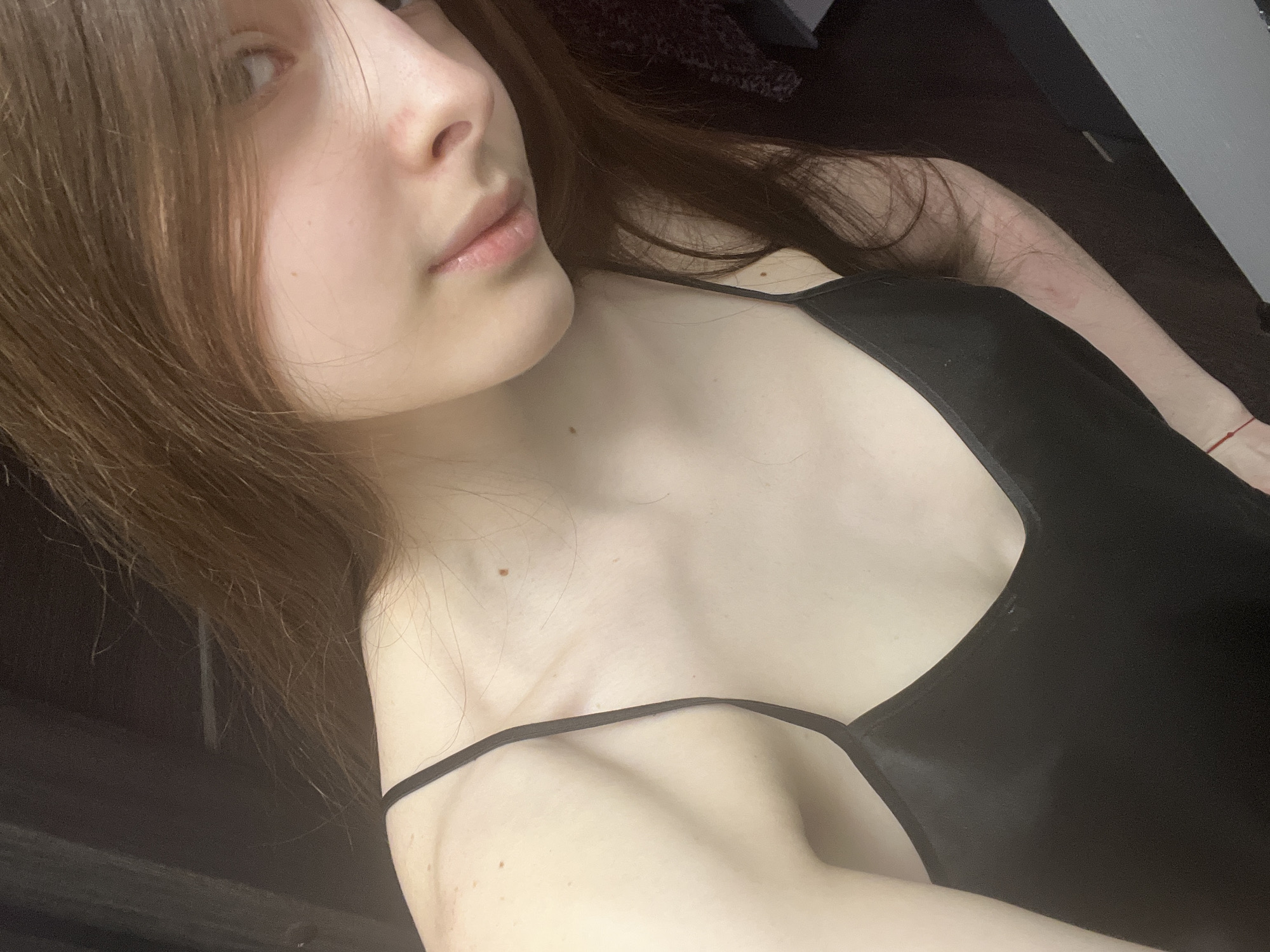 A young woman with light brown hair and fair skin is depicted in a close-up, low-angle shot. She is wearing a black camisole-style top with thin straps. Her face is partially visible, focusing on her nose, lips, and one eye. Her left arm is visible in the lower portion of the frame, while her right arm is partially visible in the upper right corner, displaying some red marks and blemishes. The background is dark and indistinct. There is no evidence to suggest the location is in Saint Petersburg, Russia.