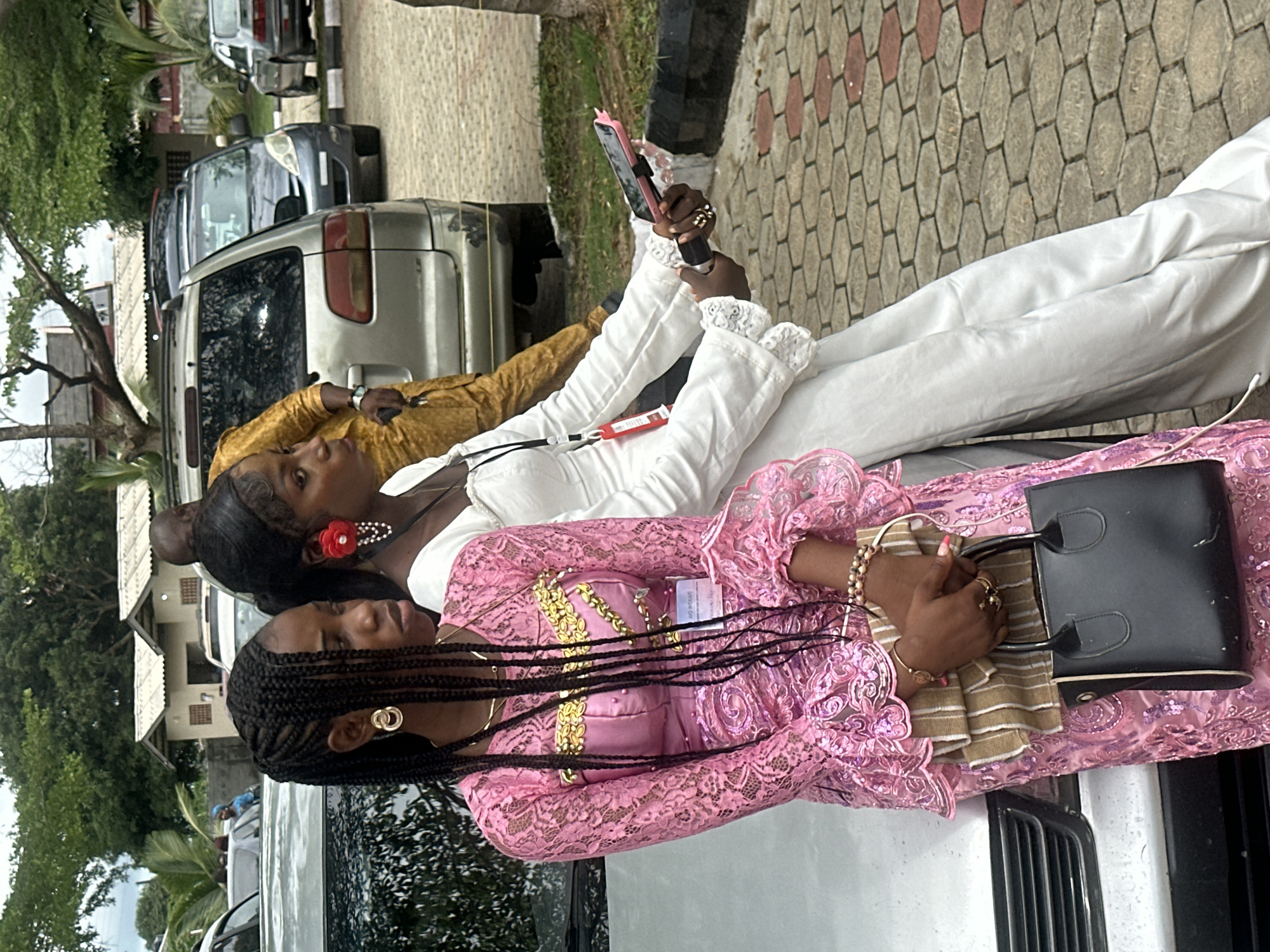 In Itoga, Nigeria, two young women are posed by the side of a silver car. The woman on the left, wearing a pink lace dress with gold embellishments, has her hair in braided extensions and is holding a black handbag. The woman on the right wears a white dress and is holding a mobile phone with a pink case, and a microphone. They appear to be at an outdoor event, with other vehicles and buildings visible in the background. The ground is paved with hexagonal tiles.