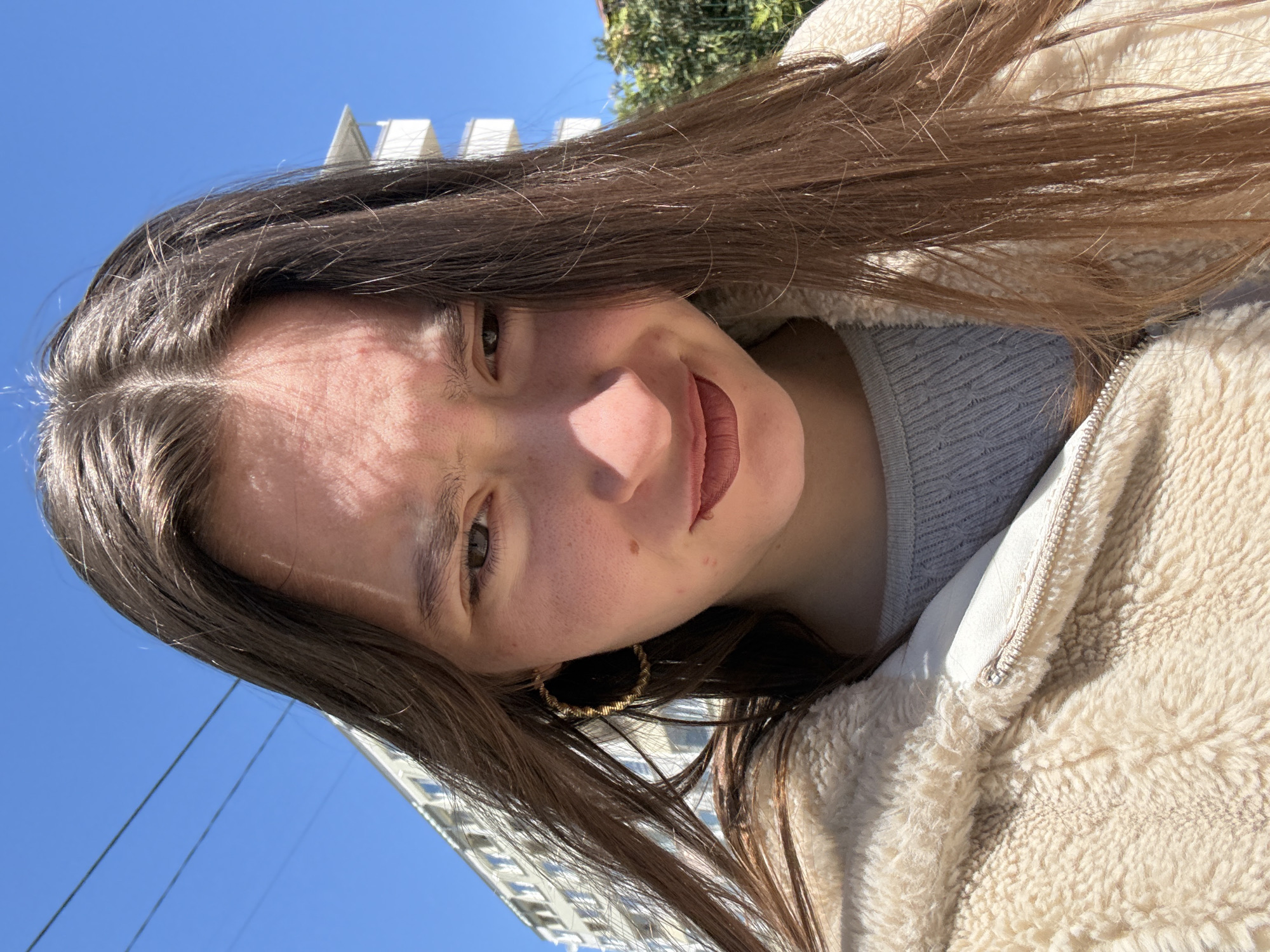 A young woman with long brown hair and hoop earrings is angled in the photo. She is wearing a light-colored, fluffy jacket over a gray sweater. The background shows a bright blue sky, a building with a white structure, trees, and electrical lines. The location is the city of Sofia, Bulgaria.