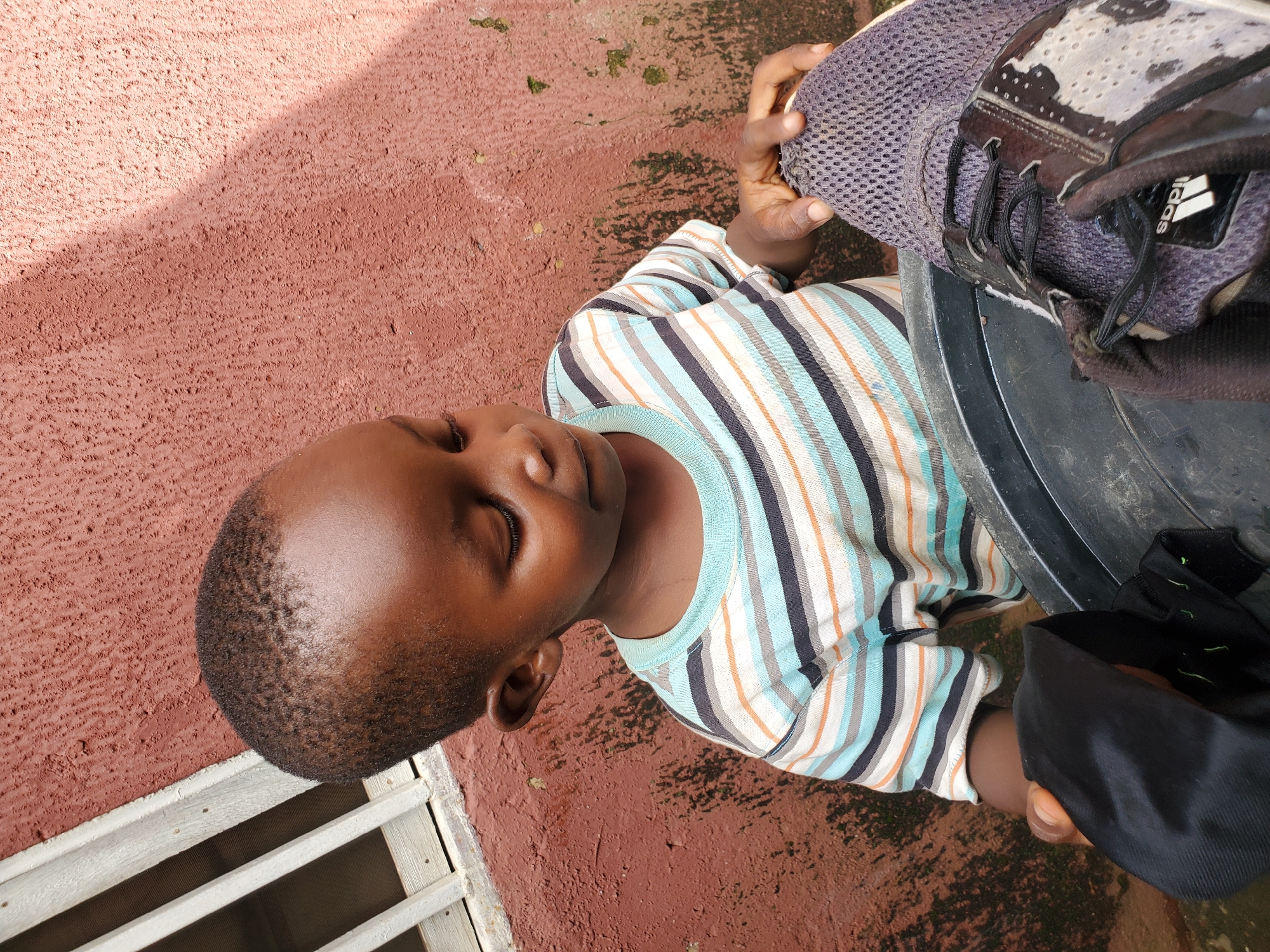 This media file features a close-up, overhead shot of a young child lying on a reddish, textured surface that appears to be an outdoor ground. The child, with their eyes closed, is wearing a horizontally striped t-shirt with bands of blue, white, orange, and black. Their head is tilted to the left, resting near a dark, lidded container, possibly a bucket or drum. A grey and black athletic shoe, with visible Adidas branding, is held by a person's hand, positioned over the child's chest and the container. The background shows the reddish ground with some patches of moss or dirt, and a sliver of a white, slatted structure, possibly a window or fence, is visible on the left. The lighting suggests it is daytime, with a shadow cast across the ground from the upper left. The overall scene evokes a sense of rest or play outdoors in a casual setting. There is no discernible text beyond the Adidas logo on the shoe.