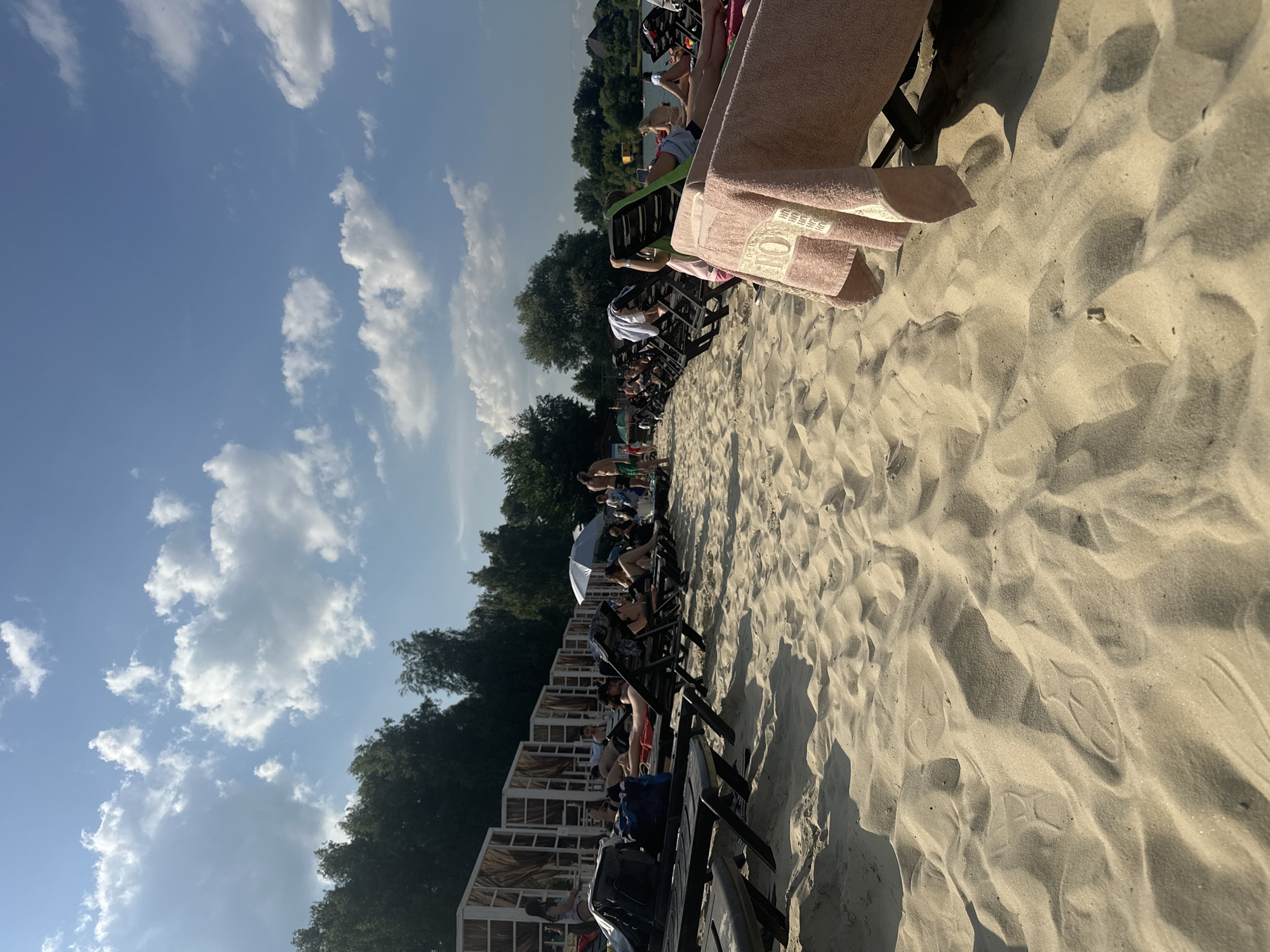 The image shows a sandy beach with several people relaxing on lounge chairs. The scene is set on a sunny day, with a bright blue sky dotted with white clouds. Trees line the background, suggesting a natural or park-like setting. Many of the people are dressed in casual beachwear, and some are facing the water, while others are engaged in conversation. A pink towel is draped over a lounge chair in the foreground, adding a splash of color to the sandy landscape. The sand itself is textured with ripples and shadows, indicating the play of sunlight. The overall mood appears to be one of leisure and relaxation. While the exact time of day is not specified, the strong sunlight and shadows suggest it is likely afternoon. The location is identified as Dal'nie Prudishchi, Russia, which aligns with the visual cues of a beach and surrounding trees. There is no visible text in the image.
