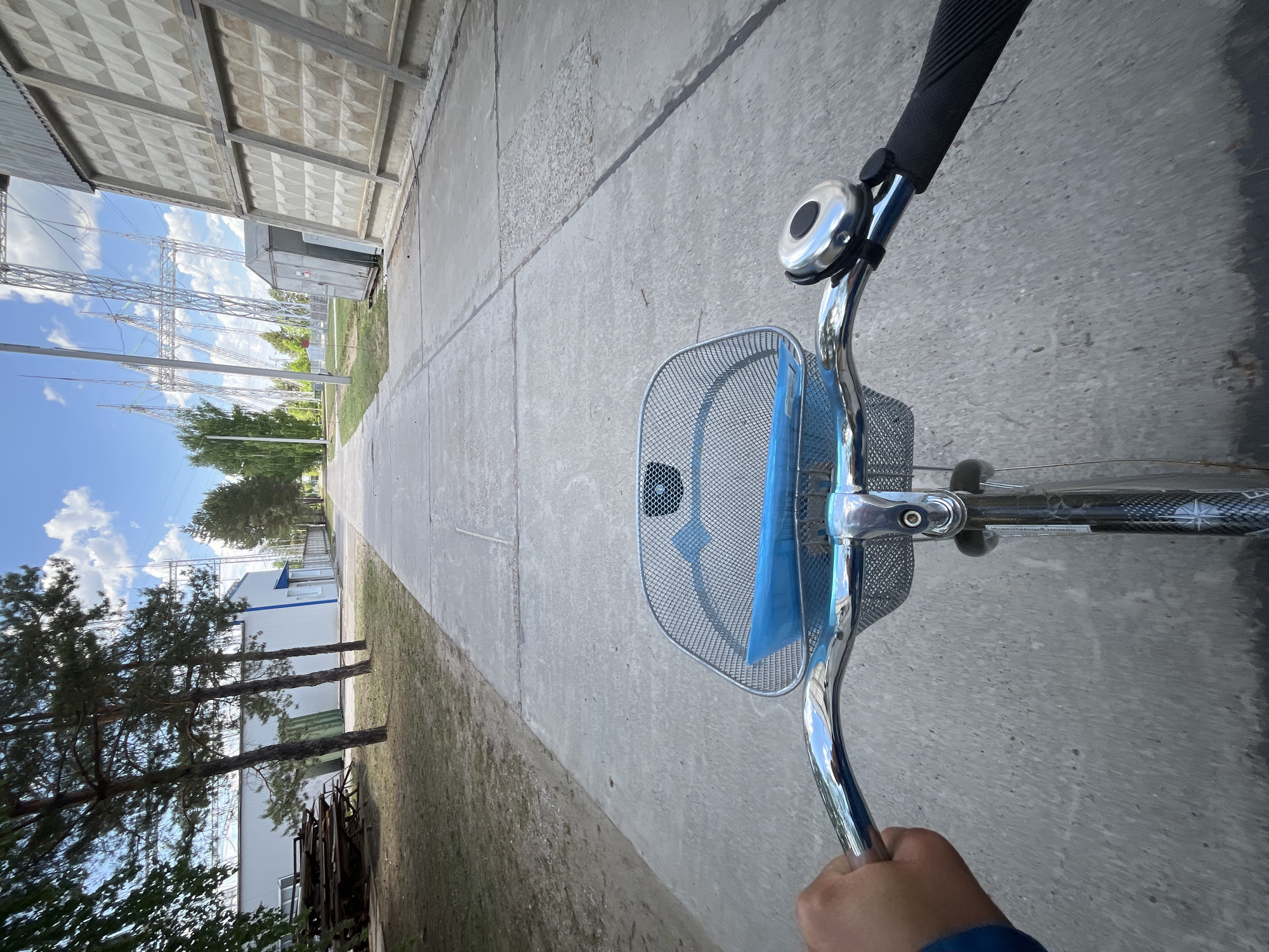 A first-person view from a bicycle is presented on a concrete path in Chaikovsky, Russia. The bicycle's handlebars, front basket, and bell are in the foreground. The basket contains a blue object. A hand clad in blue apparel grips the handlebar. The concrete path stretches into the distance, flanked by a textured concrete wall on the left and a building on the right. In the distance, electrical power pylons are visible against a partly cloudy sky. Trees line the path to the left.