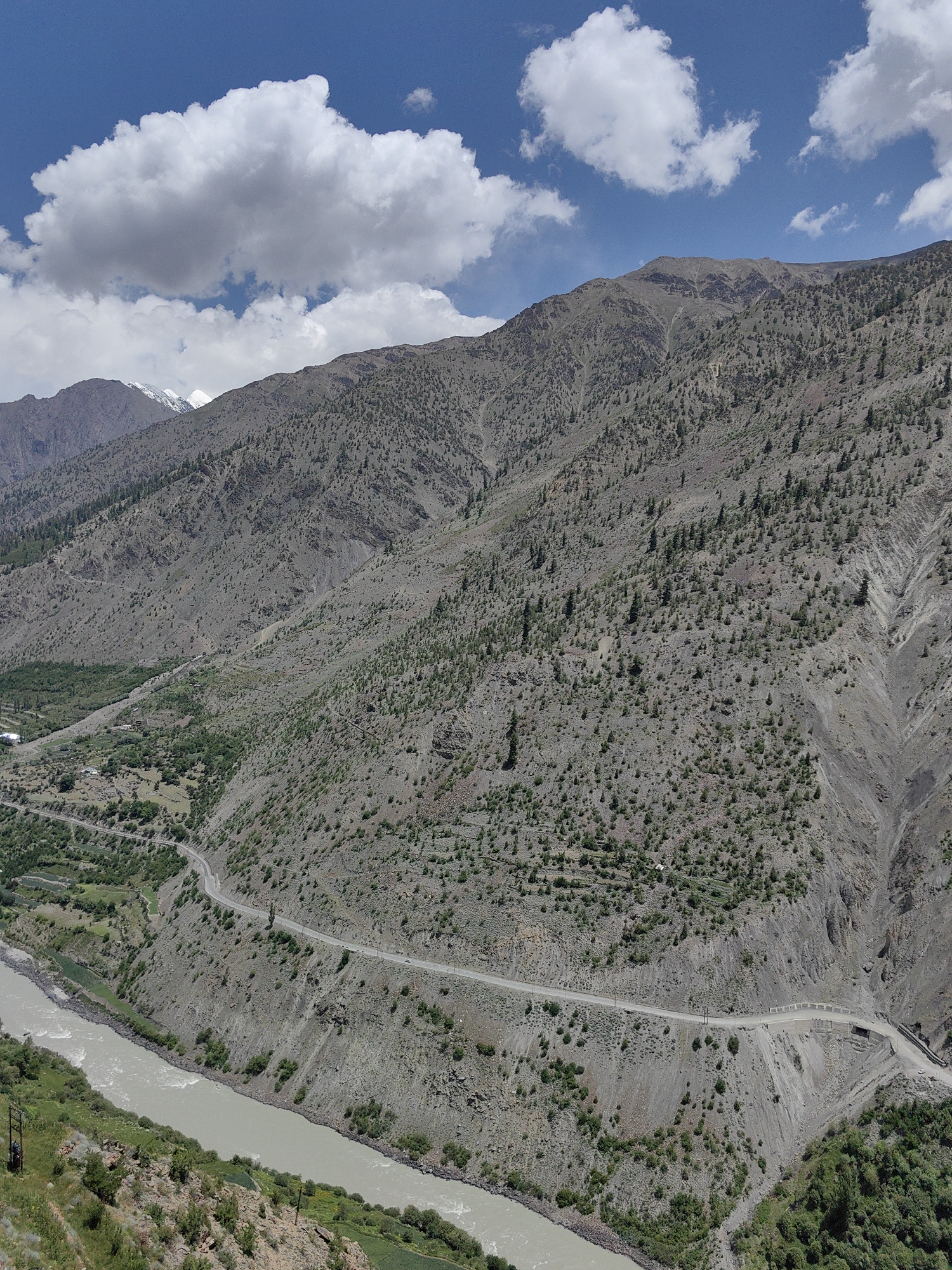 The image shows a wide, muddy river flowing through a valley with steep, rocky, and sparsely vegetated mountainsides. A winding road with guardrails snakes along the side of one mountain. Patches of green vegetation, possibly farms or small settlements, are visible in the lower left portion of the image, near the river. The sky above is blue with scattered white clouds. The scene is likely a remote, mountainous region. The mention of Kishorl, India, suggests the location is in the Indian Himalayas, known for its dramatic landscapes and river valleys.
