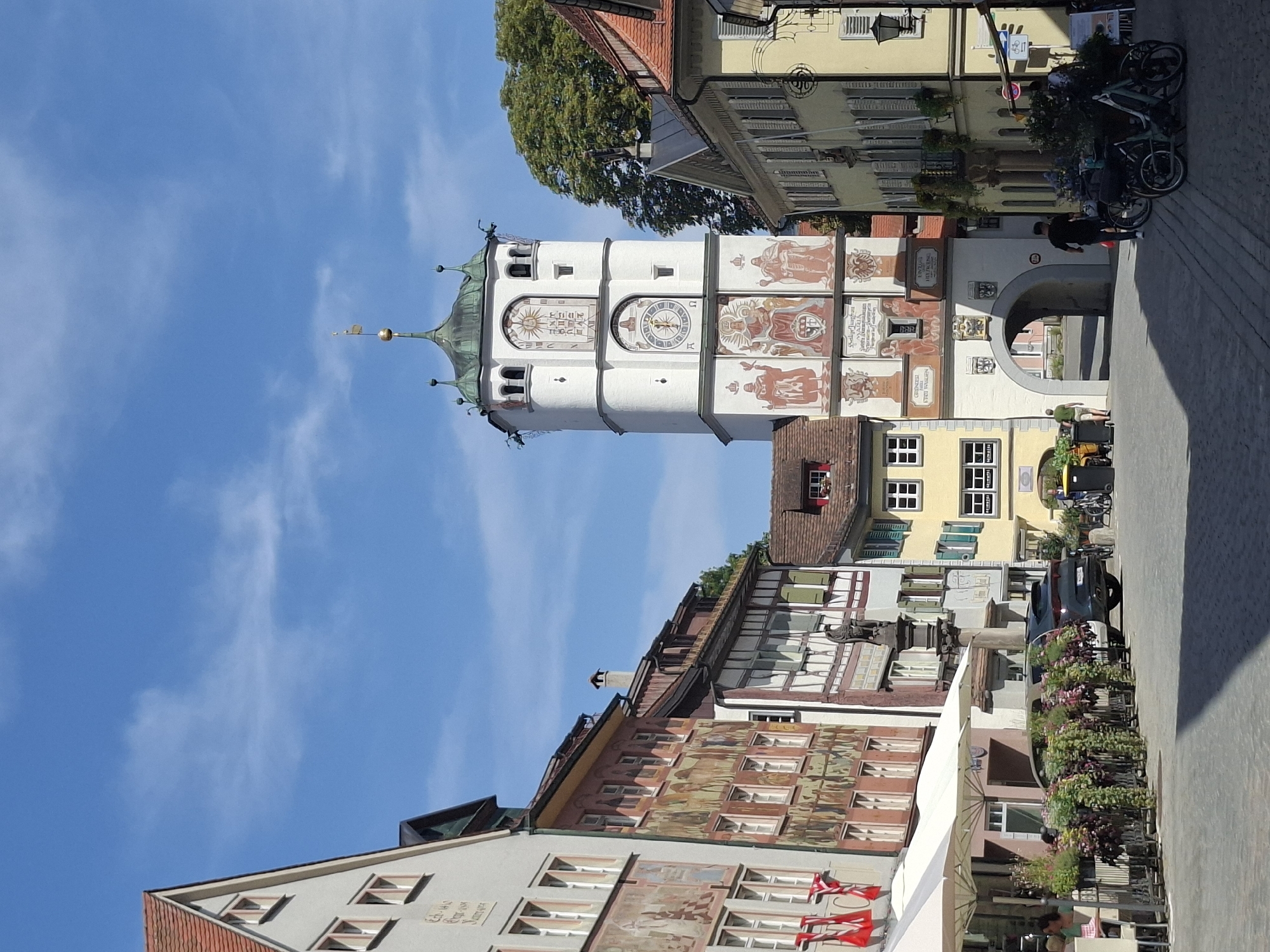 The image depicts a street scene in Wangen im Allgäu, Germany, centered on a tall, white tower with a green, multi-tiered roof and a golden spire. The tower features several painted clock faces and murals depicting figures in historical attire. An arched entryway is visible at the base of the tower.
To the left of the tower, a building with a red-tiled roof displays a mural and red banners. Adjacent to it is a building with a light-colored facade and white-framed windows. In front of these buildings, white umbrellas shade an outdoor seating area for a cafe, with several potted plants lining the front. A dark-colored car is parked along the street.
To the right of the tower, a building with a yellow facade and green shutters has a balcony with ornate railings. Another building with a red-tiled roof is partially visible. The street is paved with cobblestones.
In the foreground, several bicycles are parked. Three people are visible: one person in a dark shirt walks away from the camera towards the tower, another person in a green shirt walks towards the tower from the archway, and a third person is seated at a table under an umbrella. The sky is blue with scattered white clouds, indicating a sunny day. The overall atmosphere is that of a European town square on a pleasant day.
