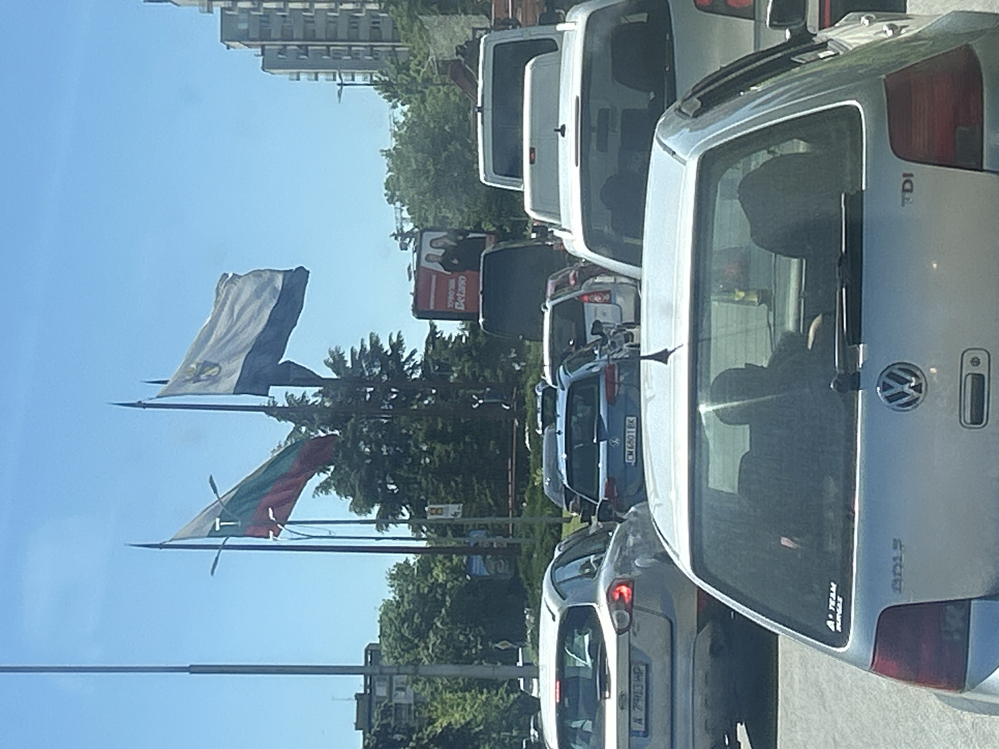 The image shows a traffic jam on a street in Burgas, Bulgaria. The back of a silver Volkswagen Golf TDI is in the foreground. Other vehicles, including vans and cars, are visible in the line of traffic. In the background are flags, including the Bulgarian flag, buildings, trees, and a billboard that partially displays an advertisement. The sky is blue.