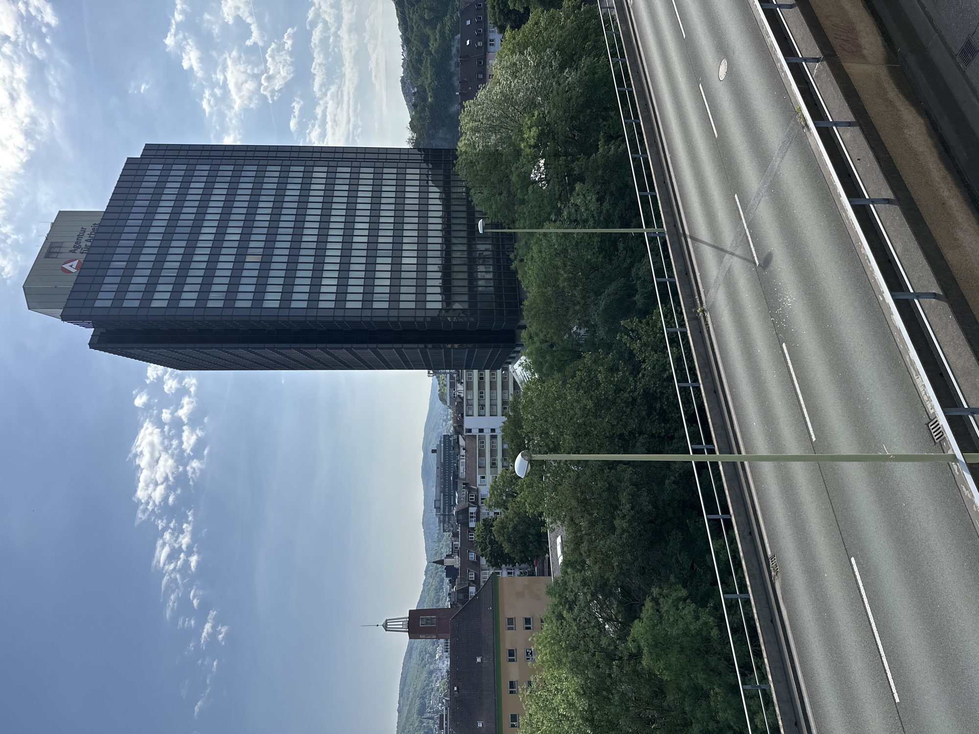 The image depicts a view of Hagen, Germany, from an elevated position, possibly a bridge or high building. A multi-story office building, identified by the logo "Agentur für Arbeit" (Employment Agency), dominates the left side of the frame. The building is of modern design with a grid pattern of windows. To the right of the building is a roadway with multiple lanes and road markings. Between the building and the road is a line of trees and the streetlights. In the background, there are buildings of varying heights and a hilly, wooded area. The sky is blue with scattered clouds. The light suggests it's daytime.