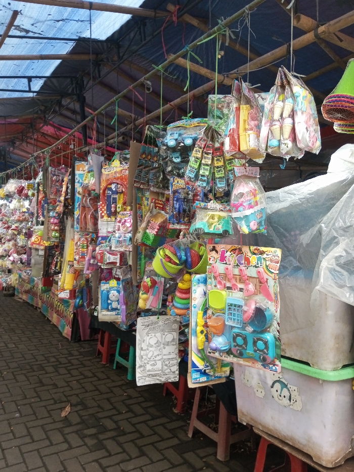 A street market is lined with stalls displaying a wide array of toys. These toys, mostly plastic and colorful, are packaged and hung from makeshift rods. Items range from toy vehicles and action figures to kitchen play sets and educational toys. The stalls are simple, often just small tables. A blue tarp covers the scene overhead, providing shade. The ground is paved with interlocking bricks.