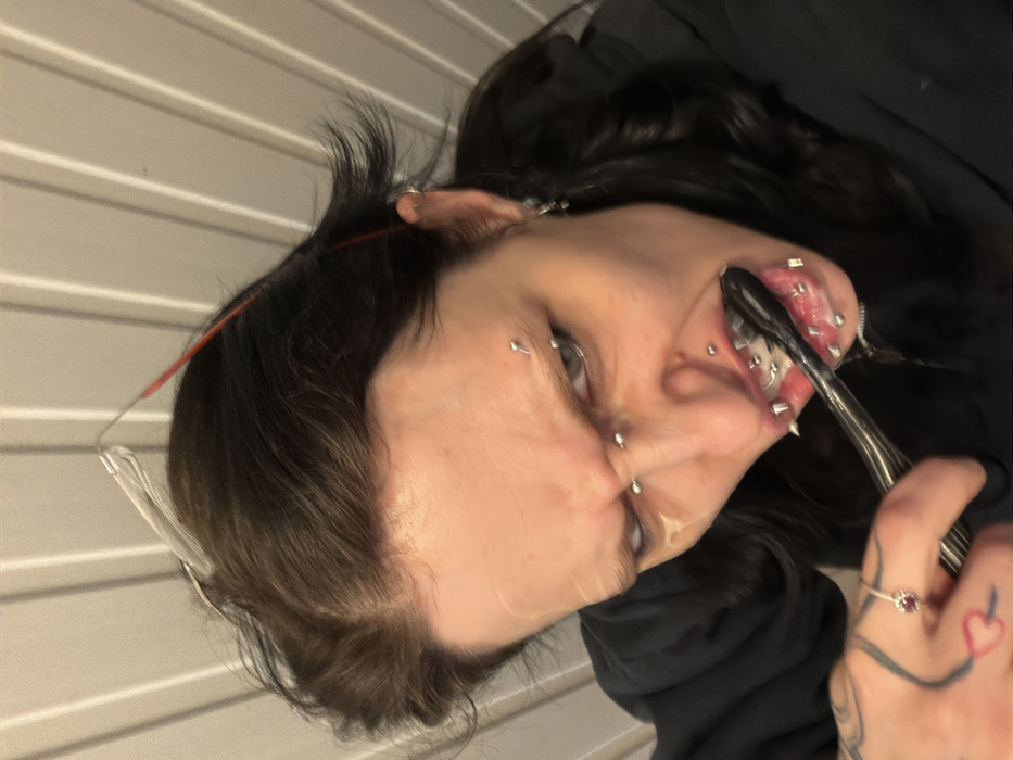 A person with numerous facial piercings, including lip and eyebrow studs, is shown from a close-up, tilted perspective. They are holding a utensil, possibly a fork, near their mouth, which is open revealing more piercings. They also have a ring on their finger. The background appears to be a textured wall. The person has dark hair and is wearing dark clothing. Part of a pair of glasses is also visible near their head.