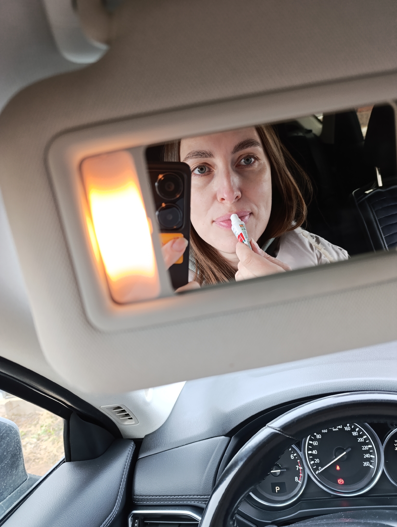The image depicts a woman applying lip balm in a car mirror, as seen from the driver's perspective. The car's dashboard, steering wheel, and instrument panel are visible, indicating the car is likely stationary with the parking brake engaged. The mirror is illuminated by a light, and a cell phone is held to capture the image in the mirror. The car's interior is visible in the reflection. While the car's interior can be assessed, information about the city of Chaikovsky, Russia is not available.