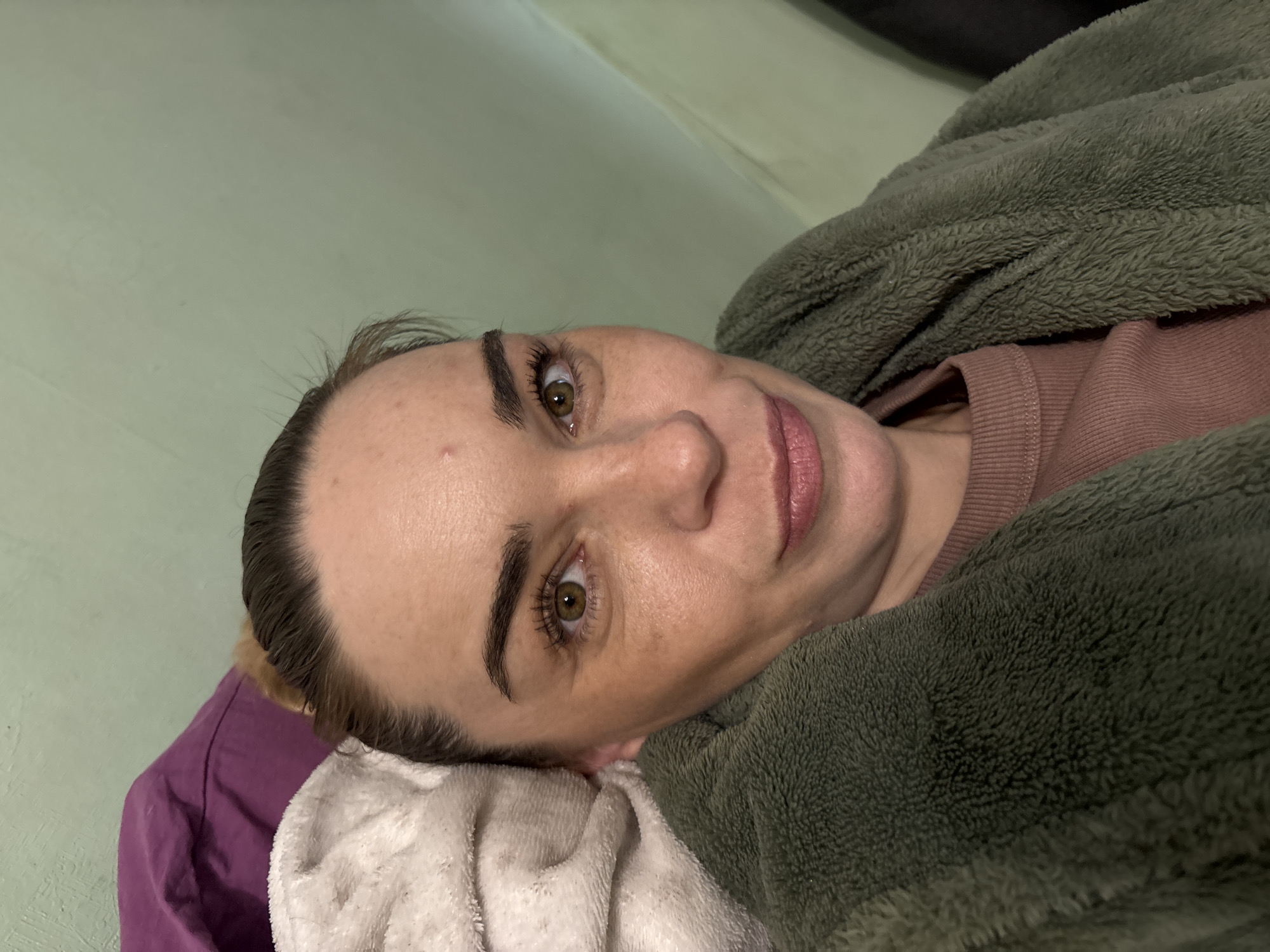 In an indoor setting with a light green wall, a woman is captured in a close-up, looking towards the camera. She has light brown hair pulled back, and her makeup includes defined eyebrows and mascara. She is wearing a pink ribbed top and a plush, olive-green robe. A purple pillow and a white, fluffy blanket are visible behind her. The lighting appears to be artificial, casting a warm glow on her skin.